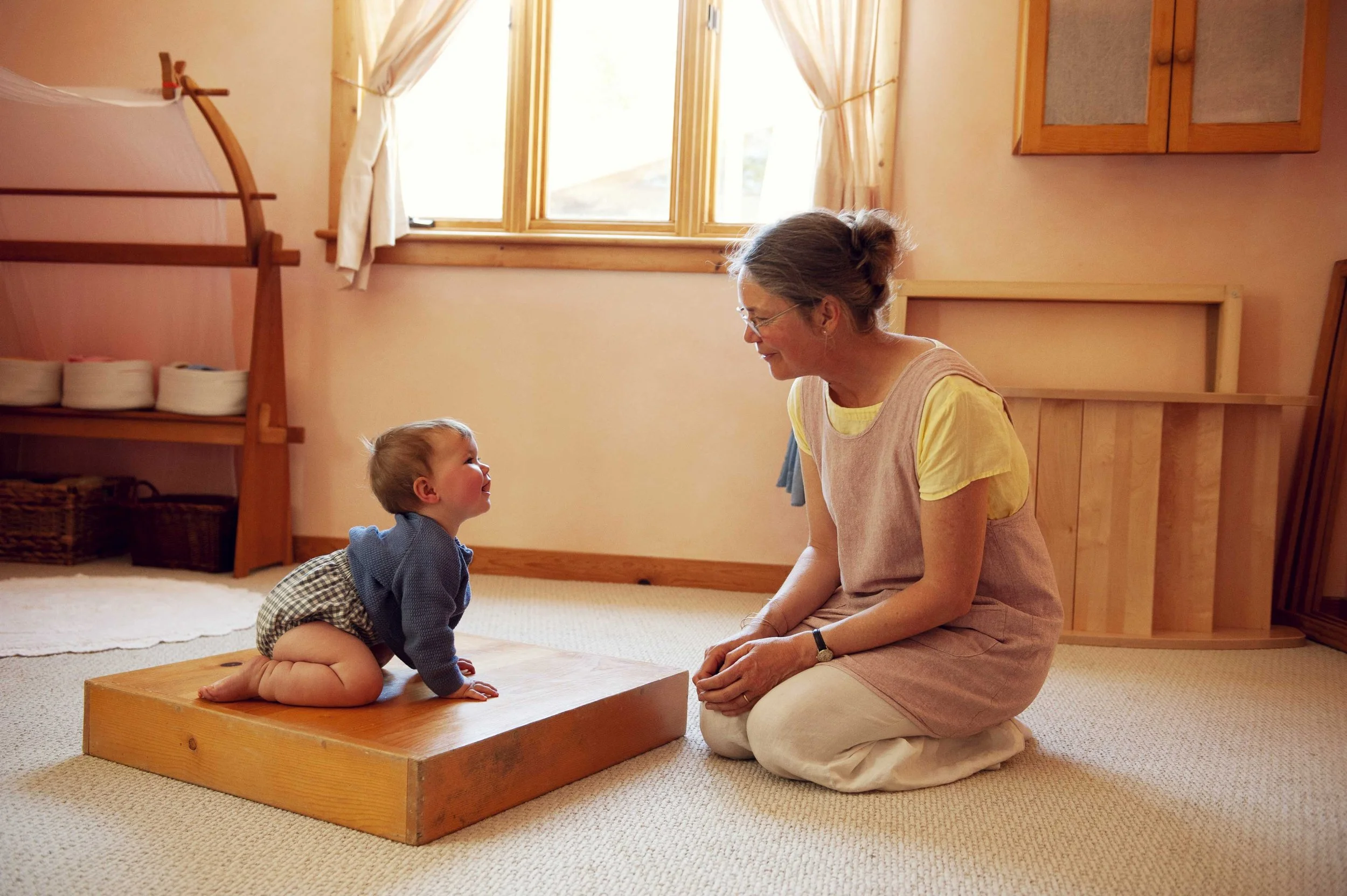 A woman and young child engaging in a Montessori-style activity in a room with warm wooden accents. The child is kneeling on a small wooden platform while the woman, sitting on her heels, smiles at the child. There is a window with curtains, a wooden