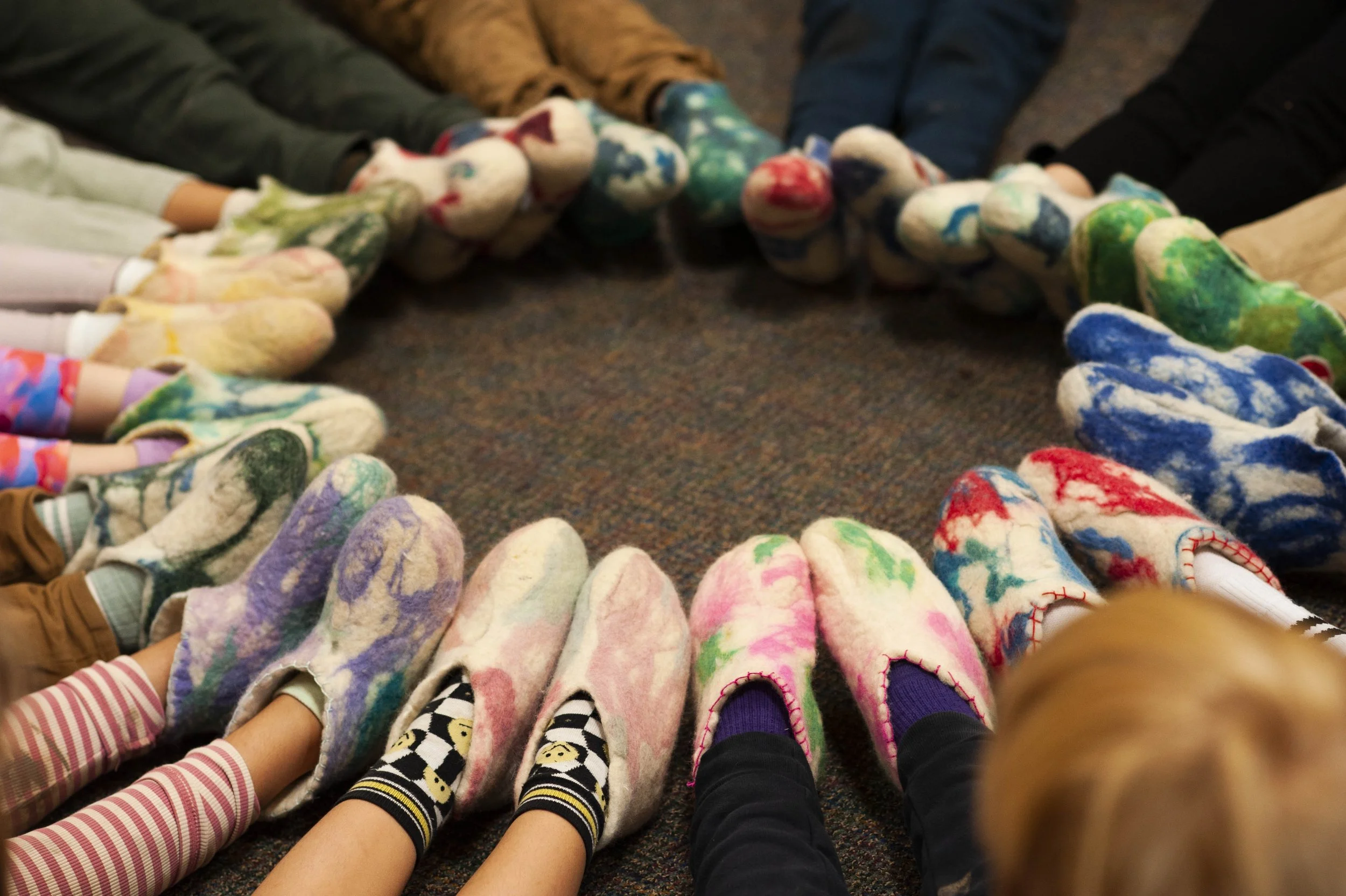 Children's feet in colorful, painted, or tie-dye slippers forming a circle on the floor.