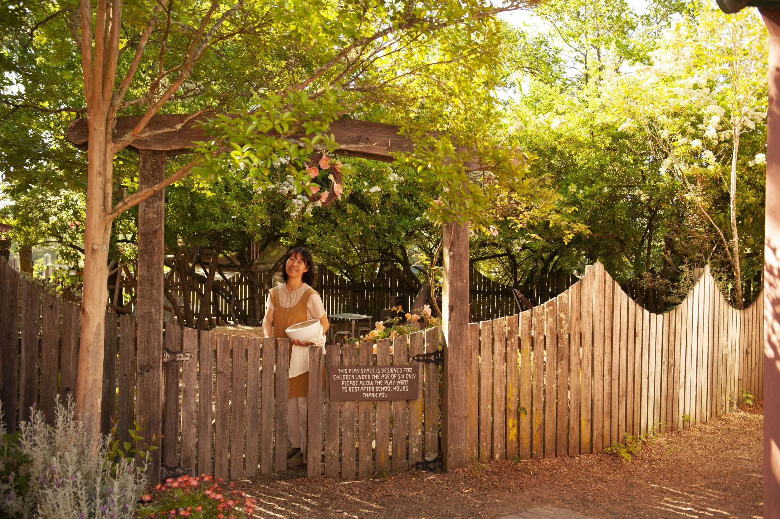 A woman in an apron holding a large white bowl standing behind a wooden fence in a garden. There are trees and shrubs, and a sign on the fence reads: 'This play space is designed for children under the age of six only. Please allow the play yard to r