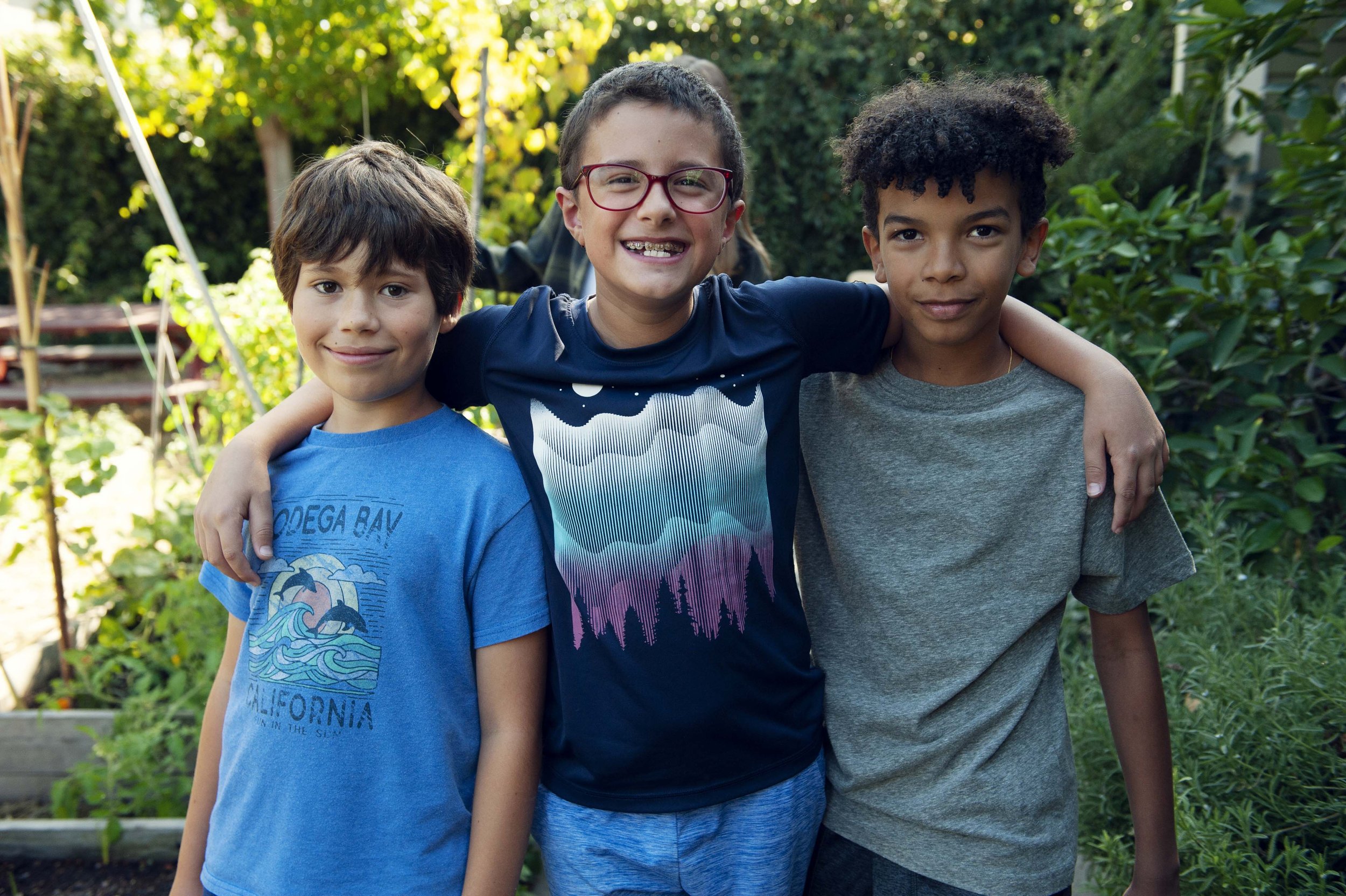 Three young boys standing outdoors with arms around each other, smiling.