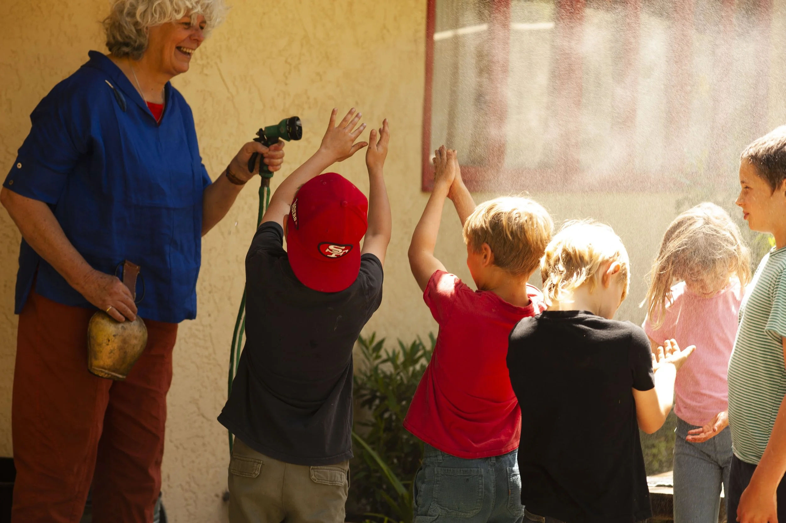 A group of children high-fiving a woman with curly gray hair, wearing a blue shirt, outdoors under a sunny sky.
