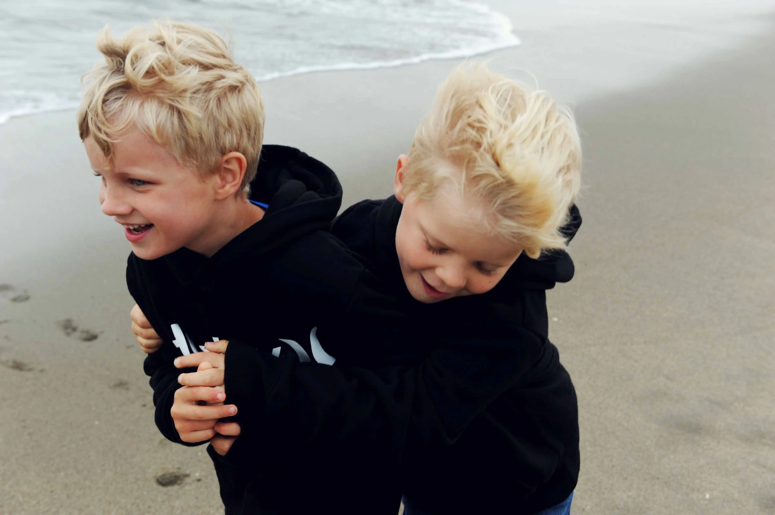 Two young boys with blonde hair playing on a beach, one piggyback on the other, near the shoreline with footprints in the sand.
