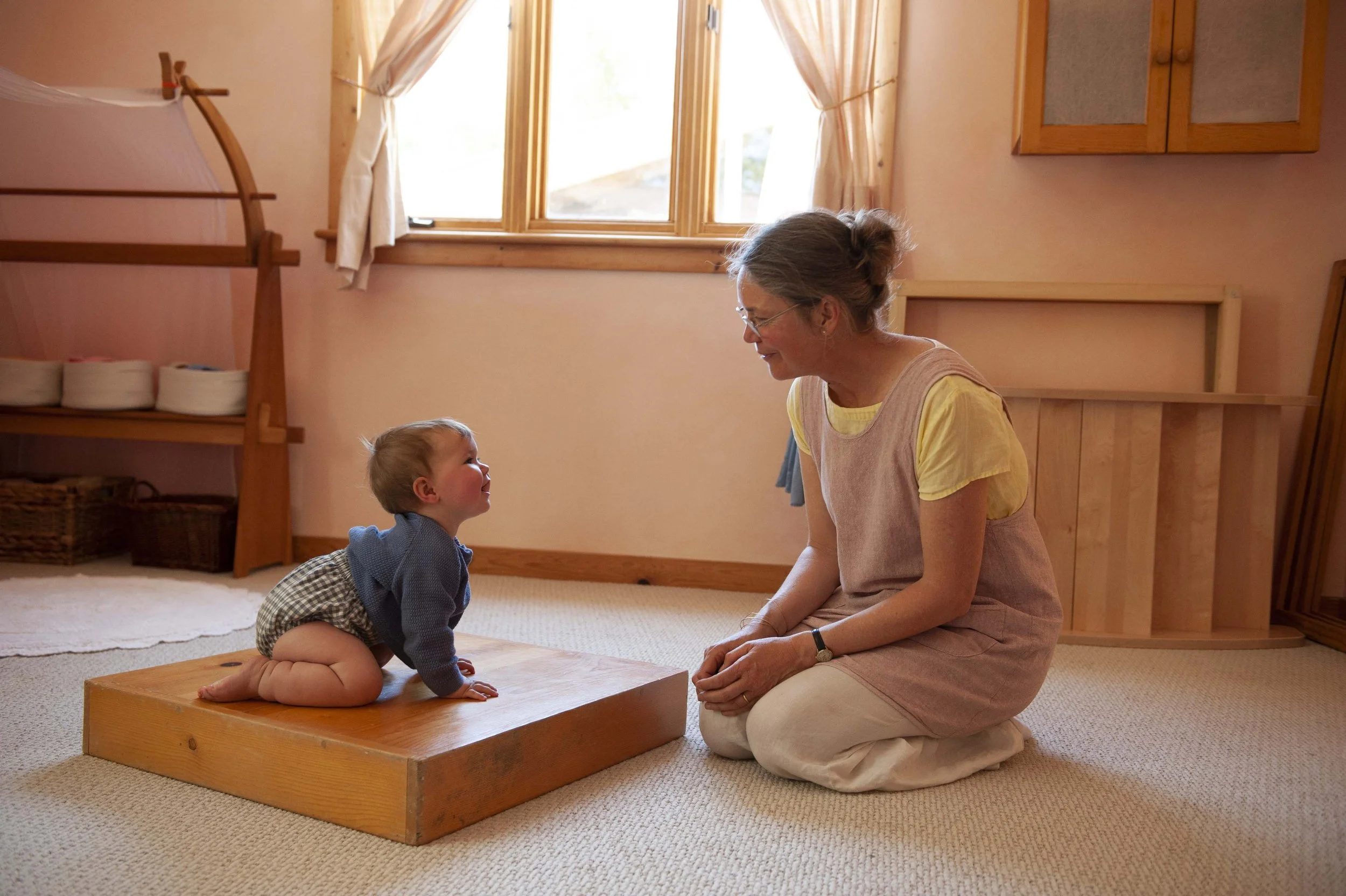An elderly woman and a young child sitting on a beige carpet in a softly lit room. The woman is kneeling and smiling at the child, who is on all fours on a wooden platform. The room has light pink walls, a window with curtains, and wooden furniture.