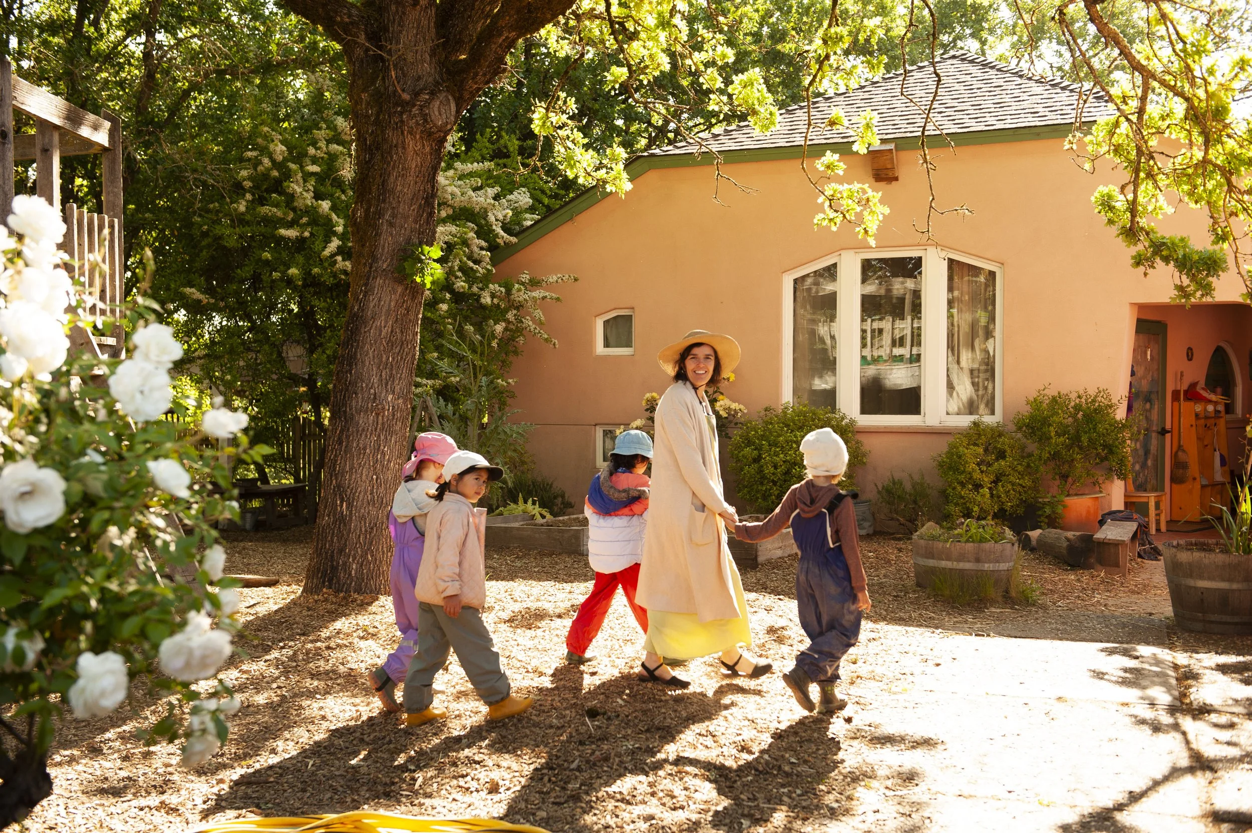 A group of children holding hands and walking in a circle outdoors, with a smiling woman in a beige coat and wide-brimmed hat, in front of a pink house with large windows and surrounded by trees and greenery.