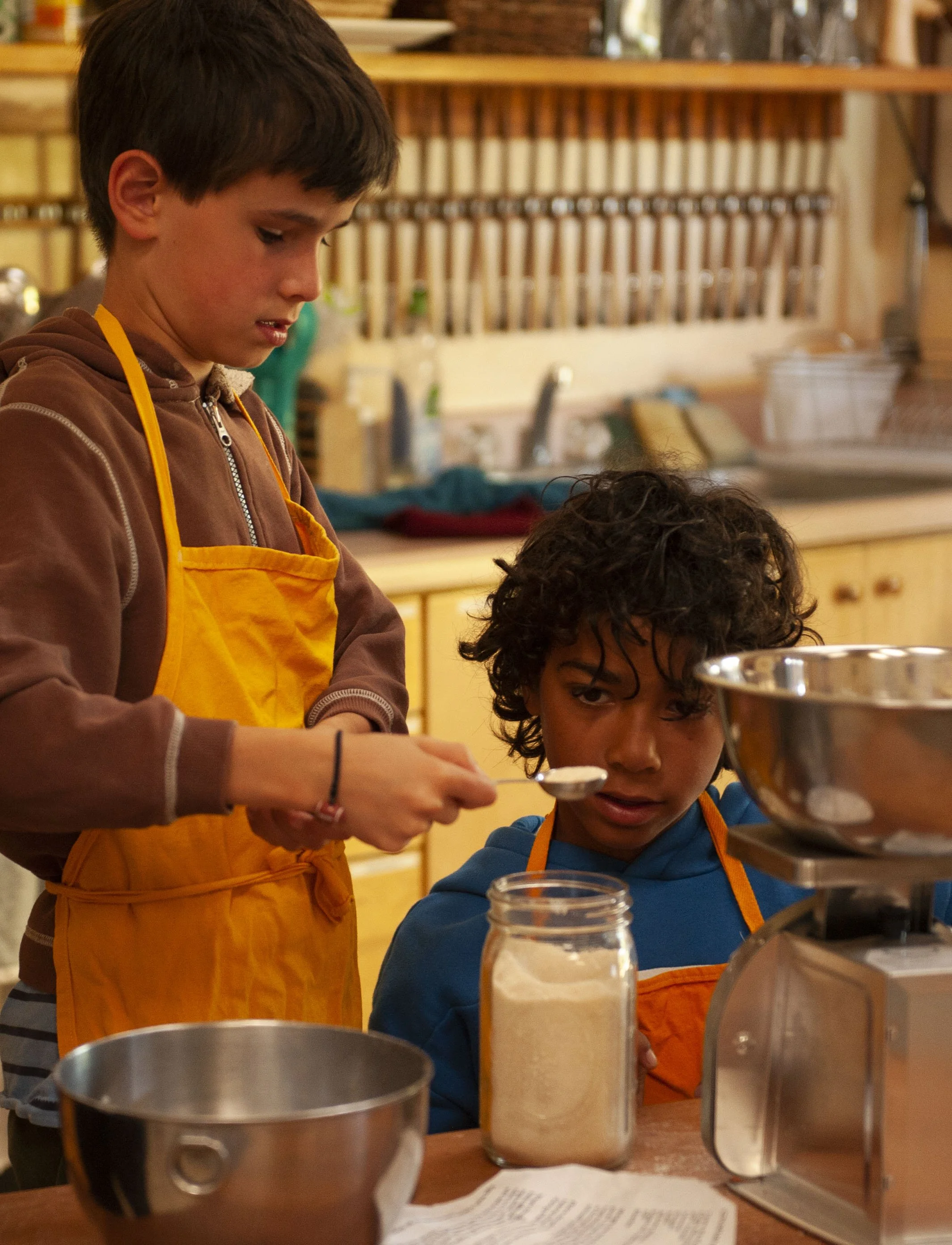 Two young boys wearing aprons are in a kitchen, one is scooping ingredients from a large jar while the other watches. The kitchen has wooden shelves with utensils and jars in the background.