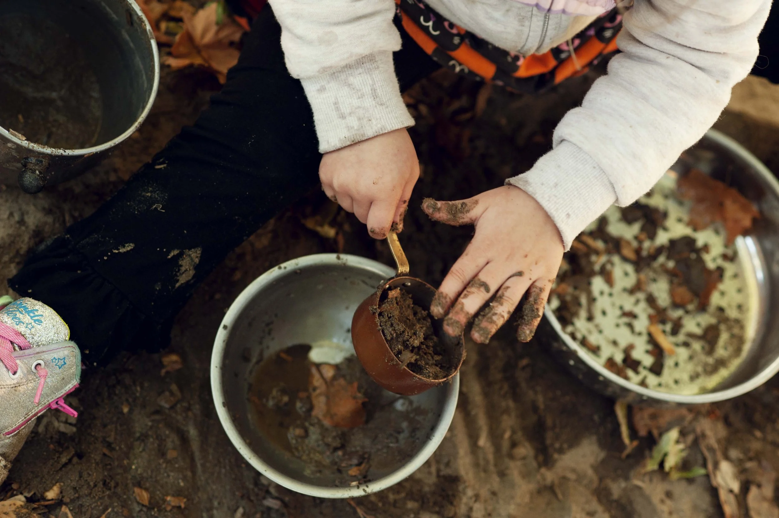 Child filling a small pot with soil from a container, surrounded by other bowls with soil and leaves, outdoors on soil ground.