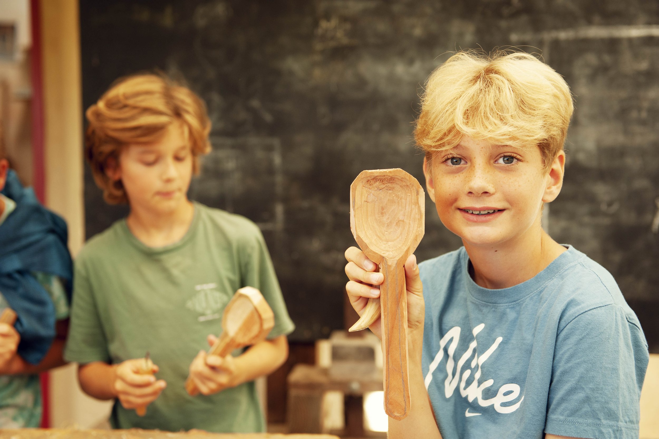 A blonde boy smiling and holding a wooden spoon in a classroom setting, with other children in the background also holding wooden utensils, in front of a black chalkboard.