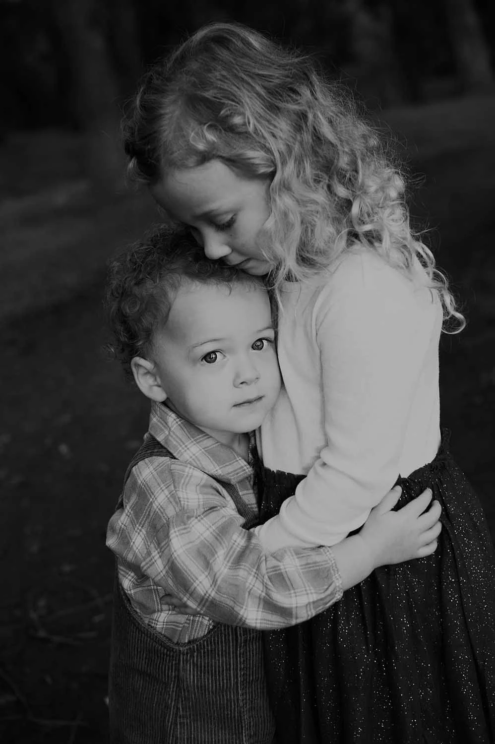 A black-and-white photo of a young girl hugging a small boy, both with serious expressions, outdoors.