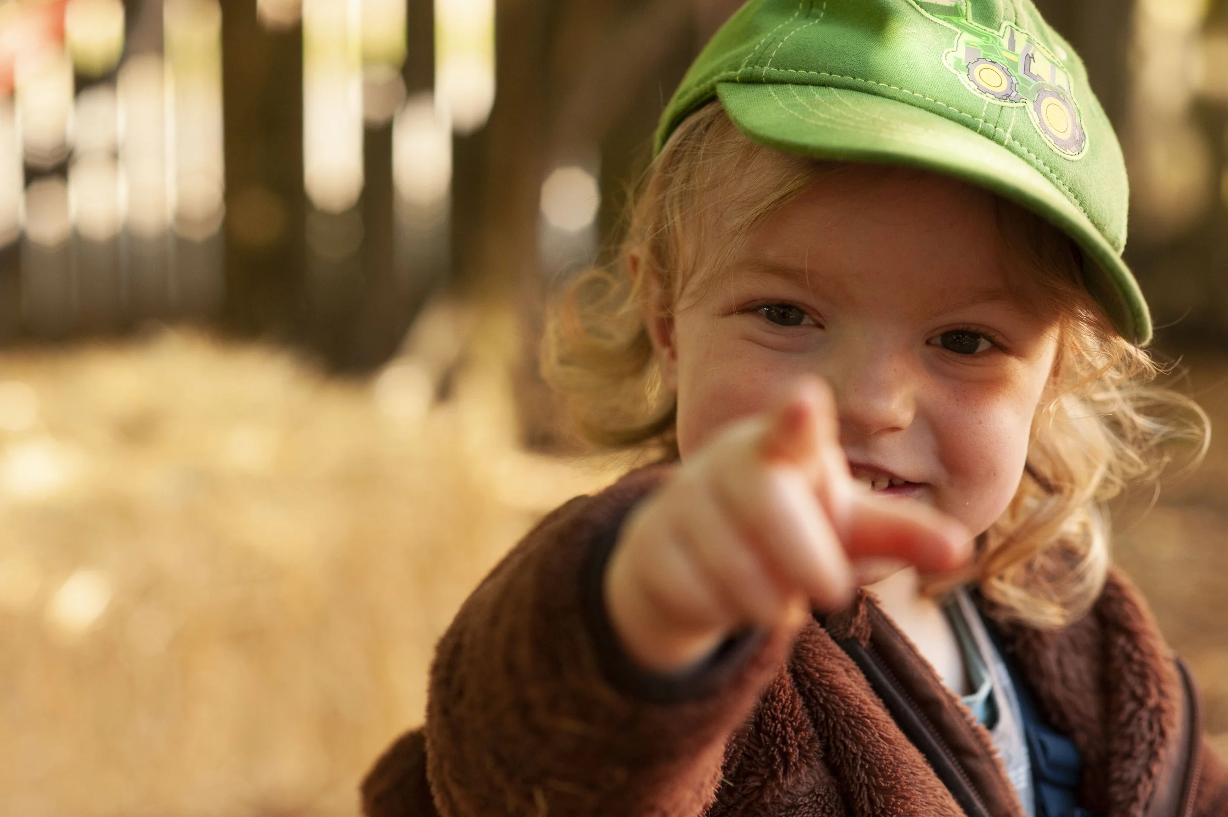 A young child with curly blonde hair wearing a green baseball cap with a tractor design, smiling and pointing towards the camera outdoors.
