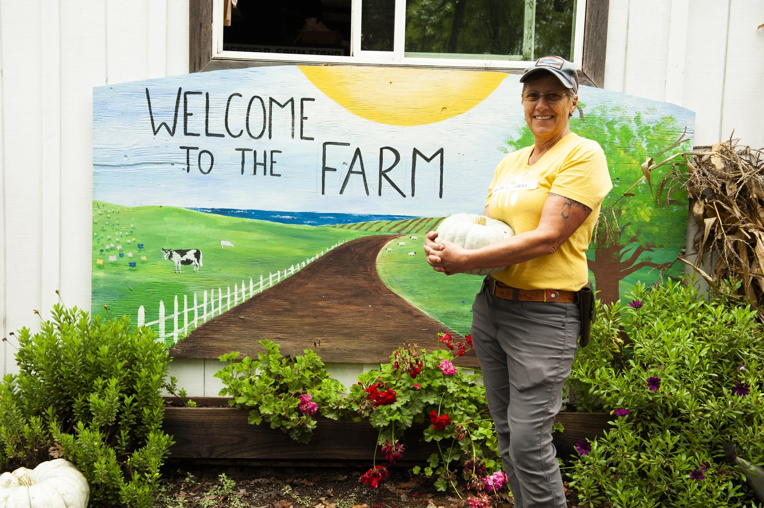 A smiling woman with gray hair, glasses, and a gray cap holding a white pumpkin stands in front of a colorful farm-themed mural that says 'Welcome to the Farm.' The mural features a green landscape, a winding road, a cow, sheep, and a large yellow su