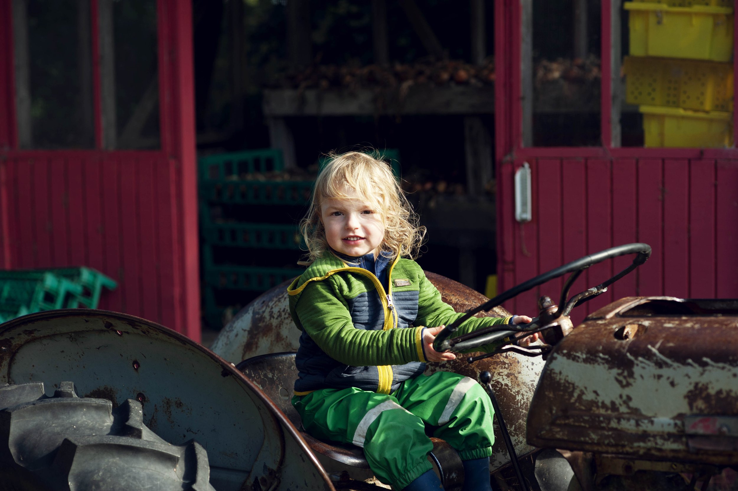Young blonde girl in green and navy outdoor clothing sitting on a rusty tractor, holding the steering wheel, with a red barn and stacked yellow crates in the background.