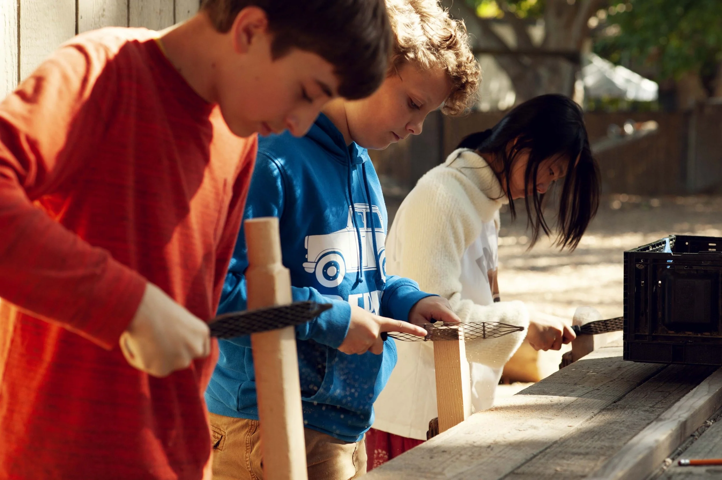 Three children focused on woodworking projects outdoors, with tools and wood pieces on a wooden workbench.