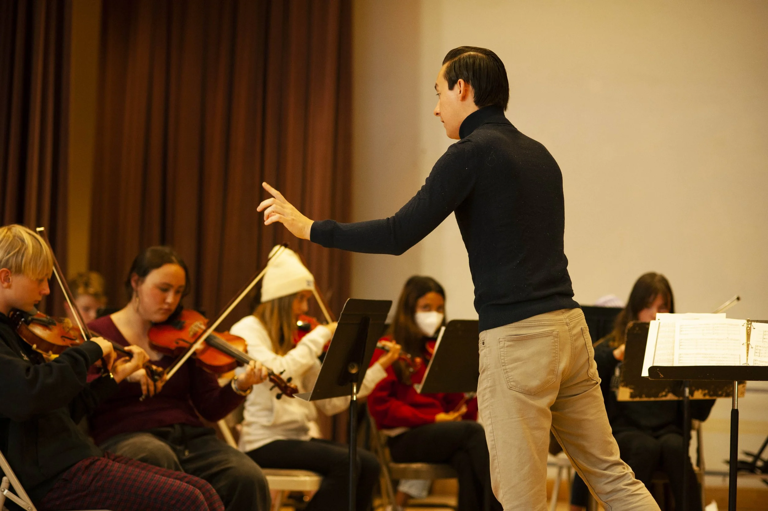 Orchestra conductor leading young violinists during rehearsal, with music stands and sheet music, in a room with brown curtains.