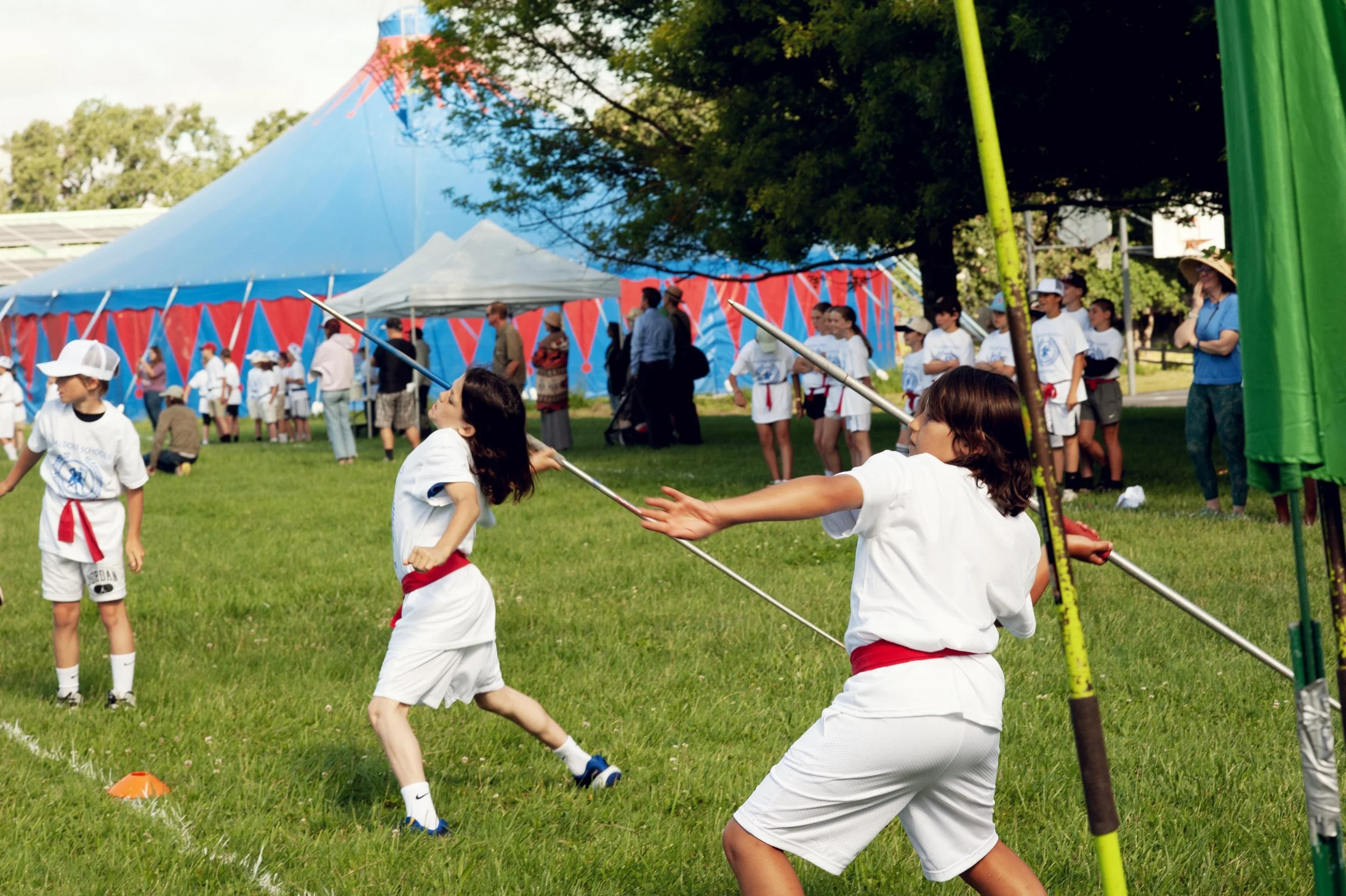 Children participating in a martial arts demonstration at an outdoor event, with a large blue and red circus tent in the background and spectators watching.