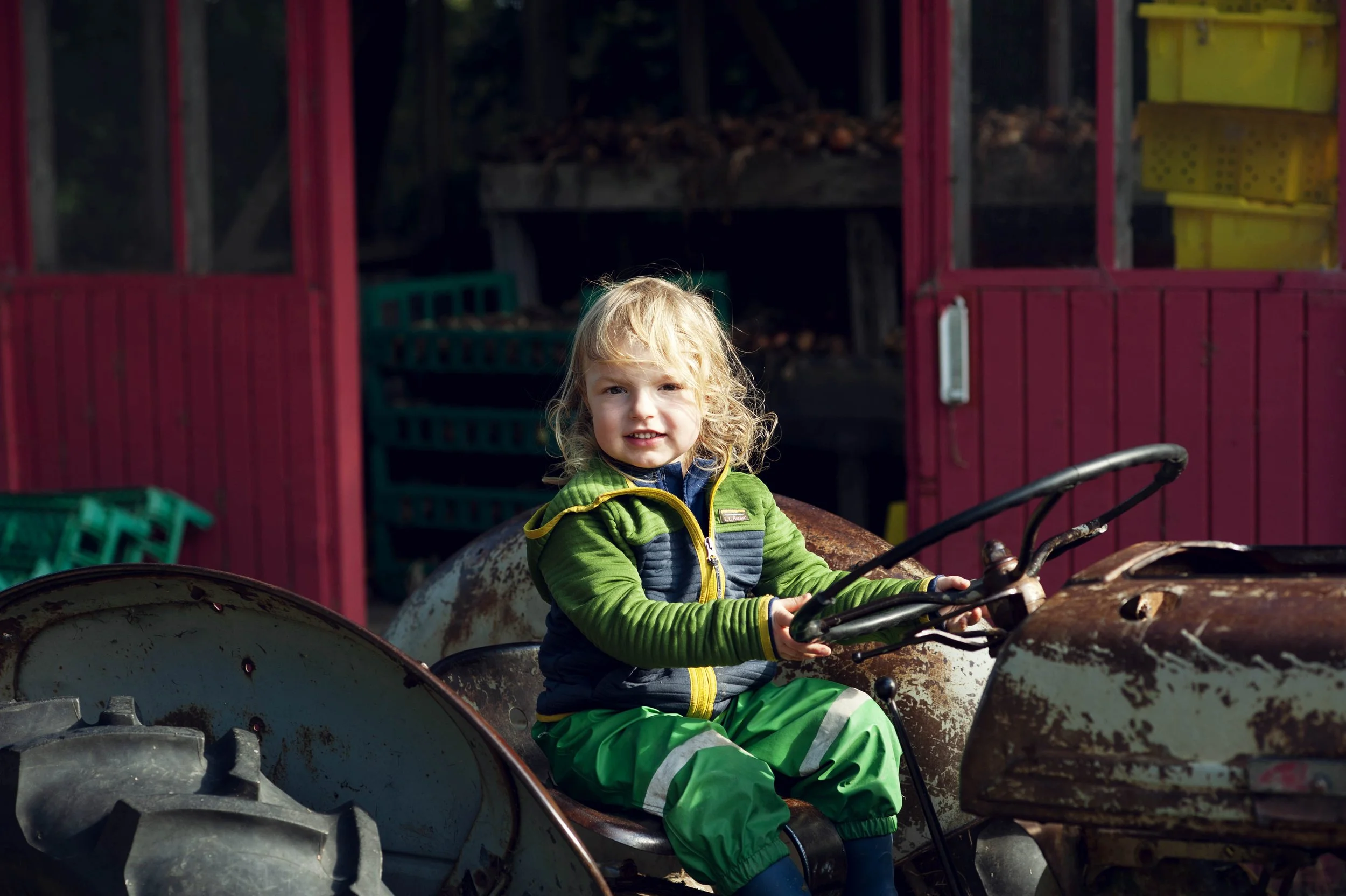 A young child with blonde curly hair and a green jacket sitting on a vintage tractor. The background features a red barn and some yellow storage bins.