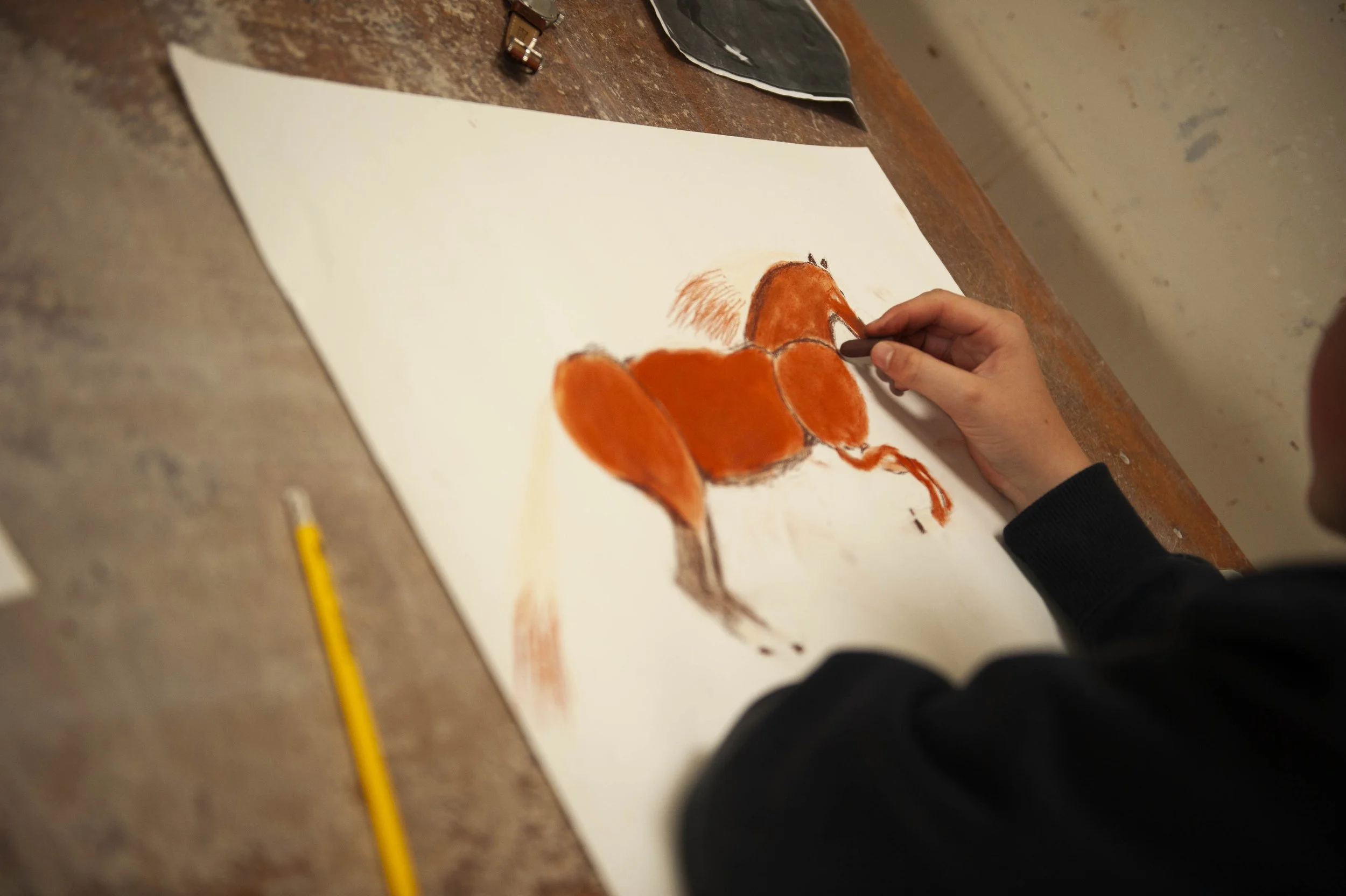 A person is drawing an orange-colored fox on a large white sheet of paper using charcoal or a pencil, on a wooden table, with art supplies nearby.