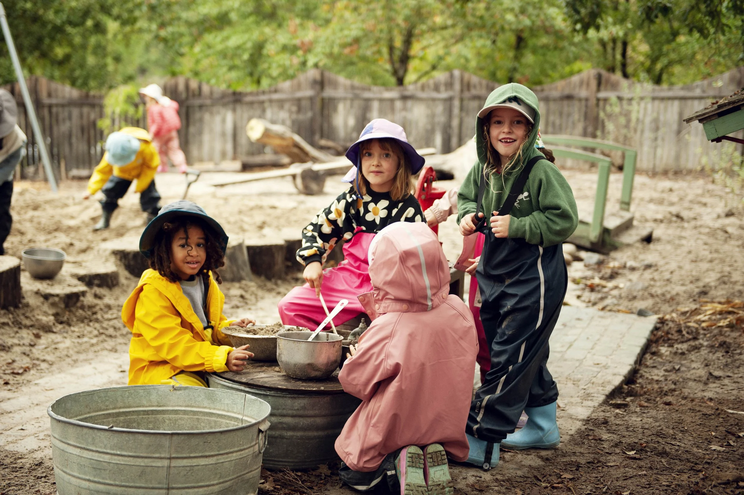 Children dressed in colorful rain gear playing outside in a sandy area with wooden logs and fence in the background.