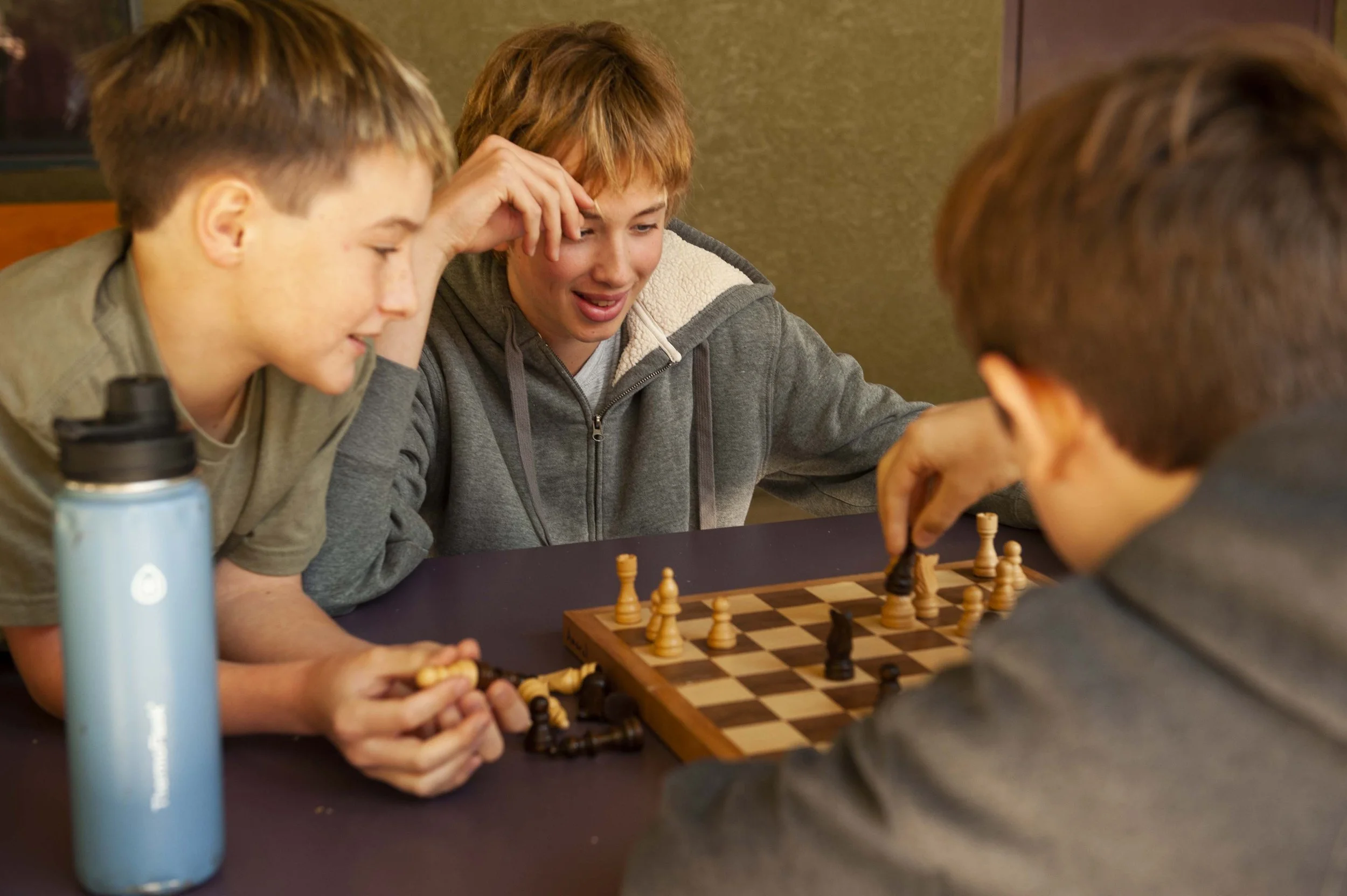 Three boys playing chess at a table, with a water bottle nearby.