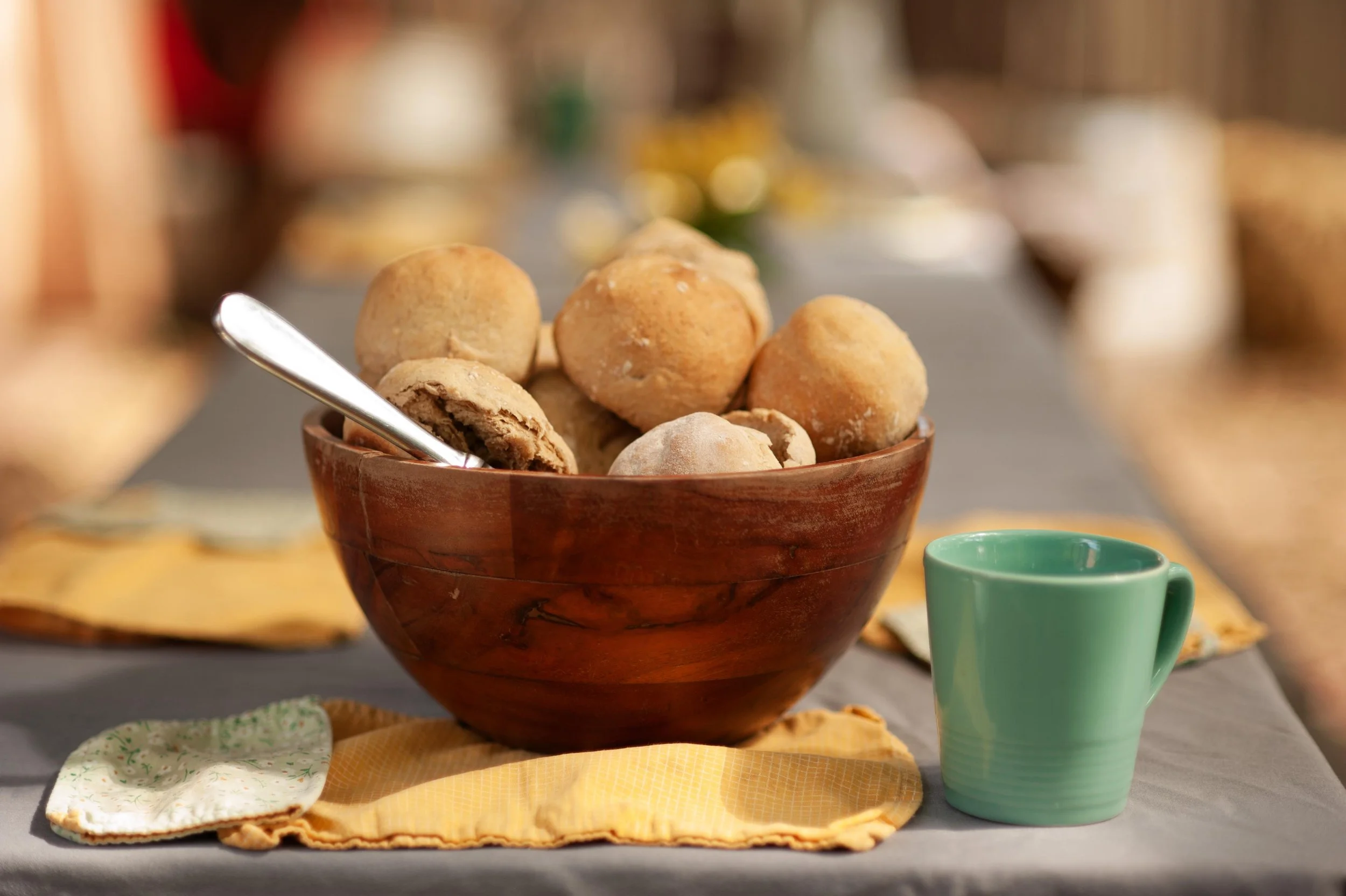 A wooden bowl filled with round, golden-brown cookies on a table with a yellow napkin, a small green mug nearby, and blurred background of more baked goods and decorations.