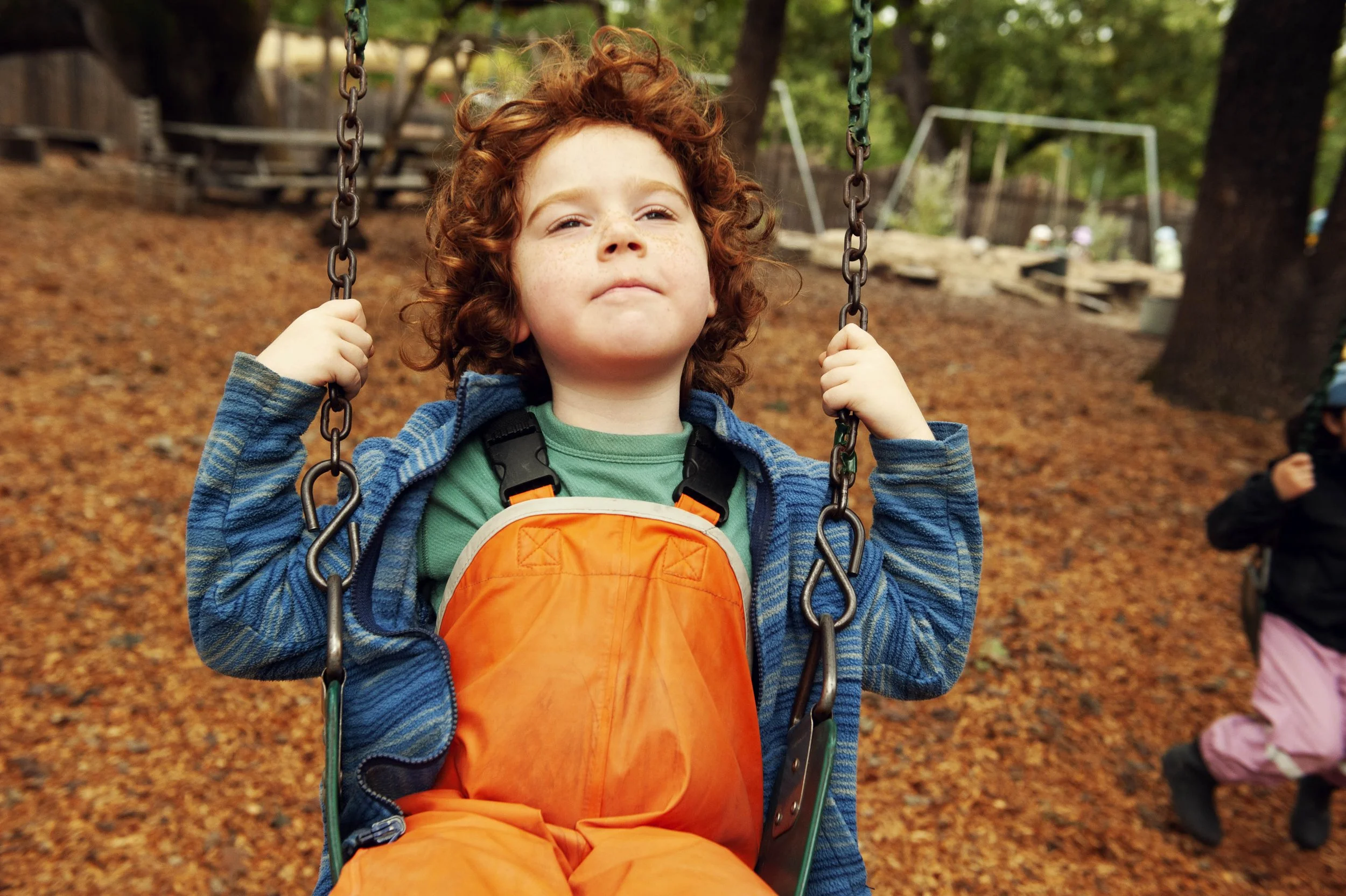 A young boy with curly red hair and freckles sitting on a swing in a park, wearing an orange jacket and a green shirt. The background shows a playground covered with fallen autumn leaves, with trees and blurred playground equipment.