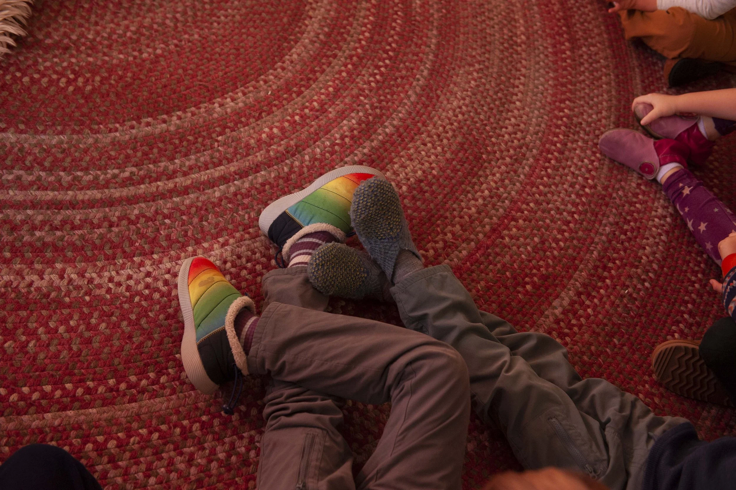 Children sitting on a red patterned carpet, with their legs crossed or extended, wearing colorful rainbow shoes and socks with various patterns.