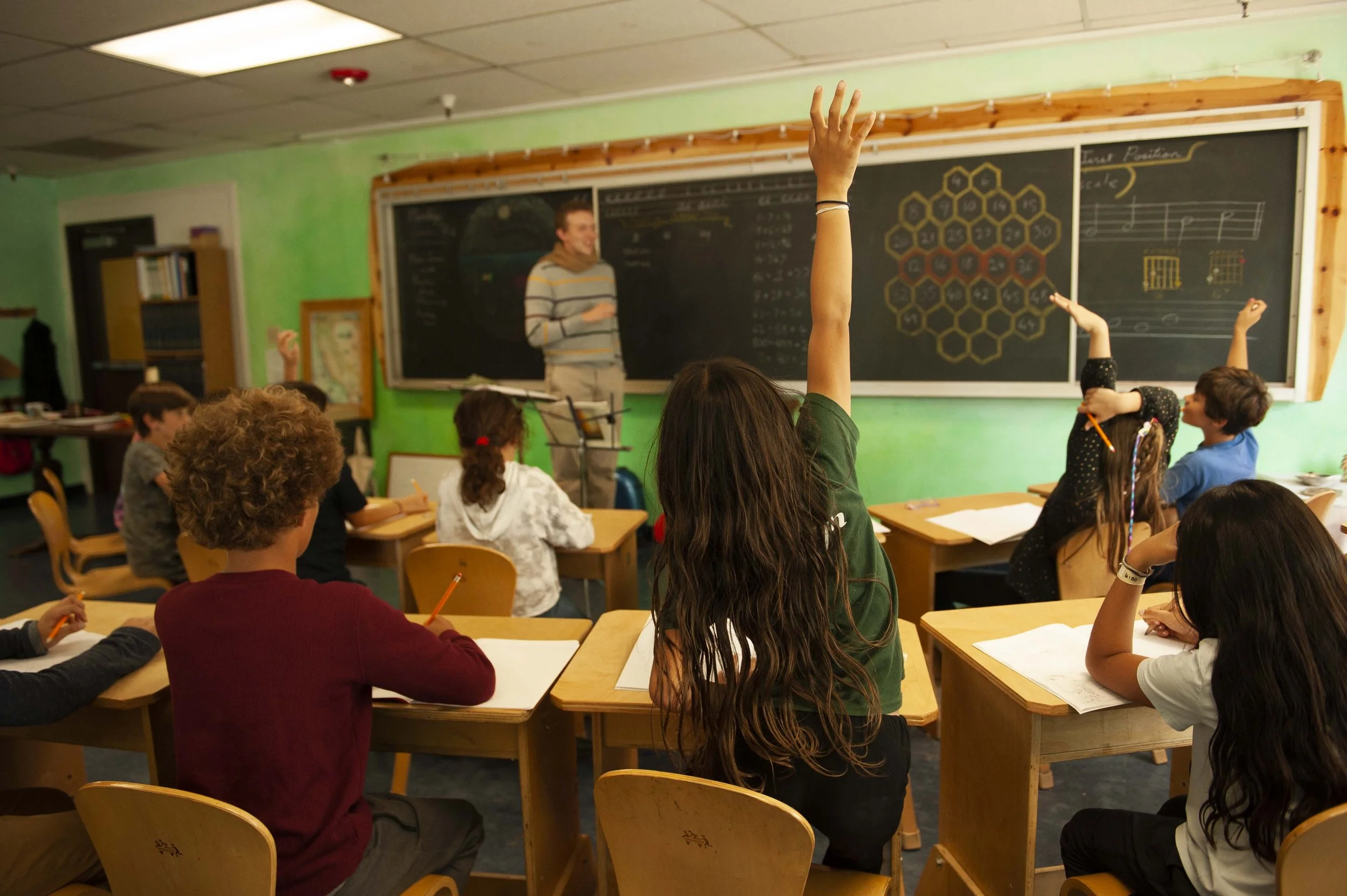 A classroom with children sitting at desks, one girl raising her hand, another girl with a headband, and a teacher at the blackboard.