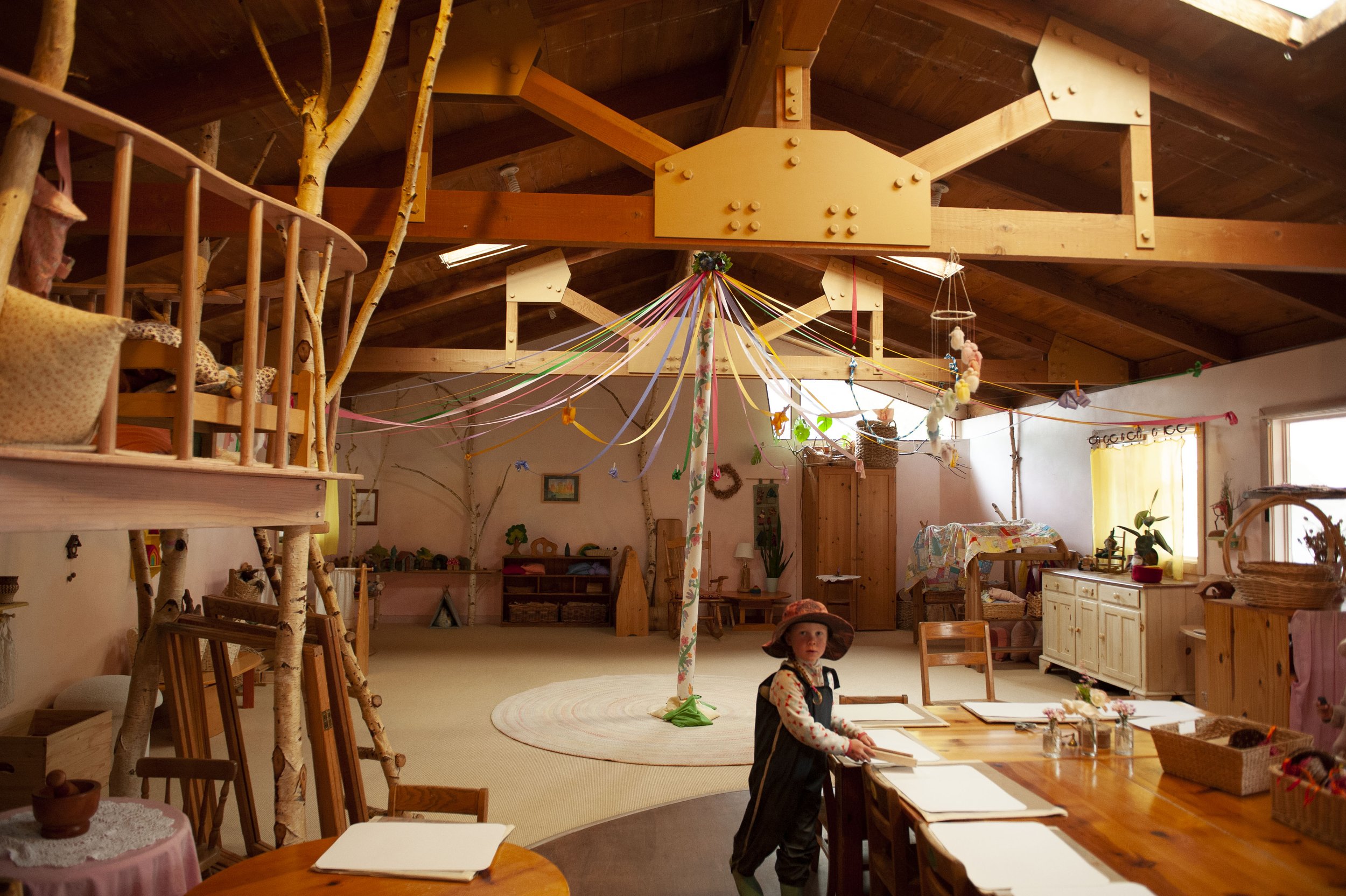 Inside a cozy, wood-paneled room decorated with colorful ribbons and crafts, with a toddler sitting at a table and a young girl standing nearby.
