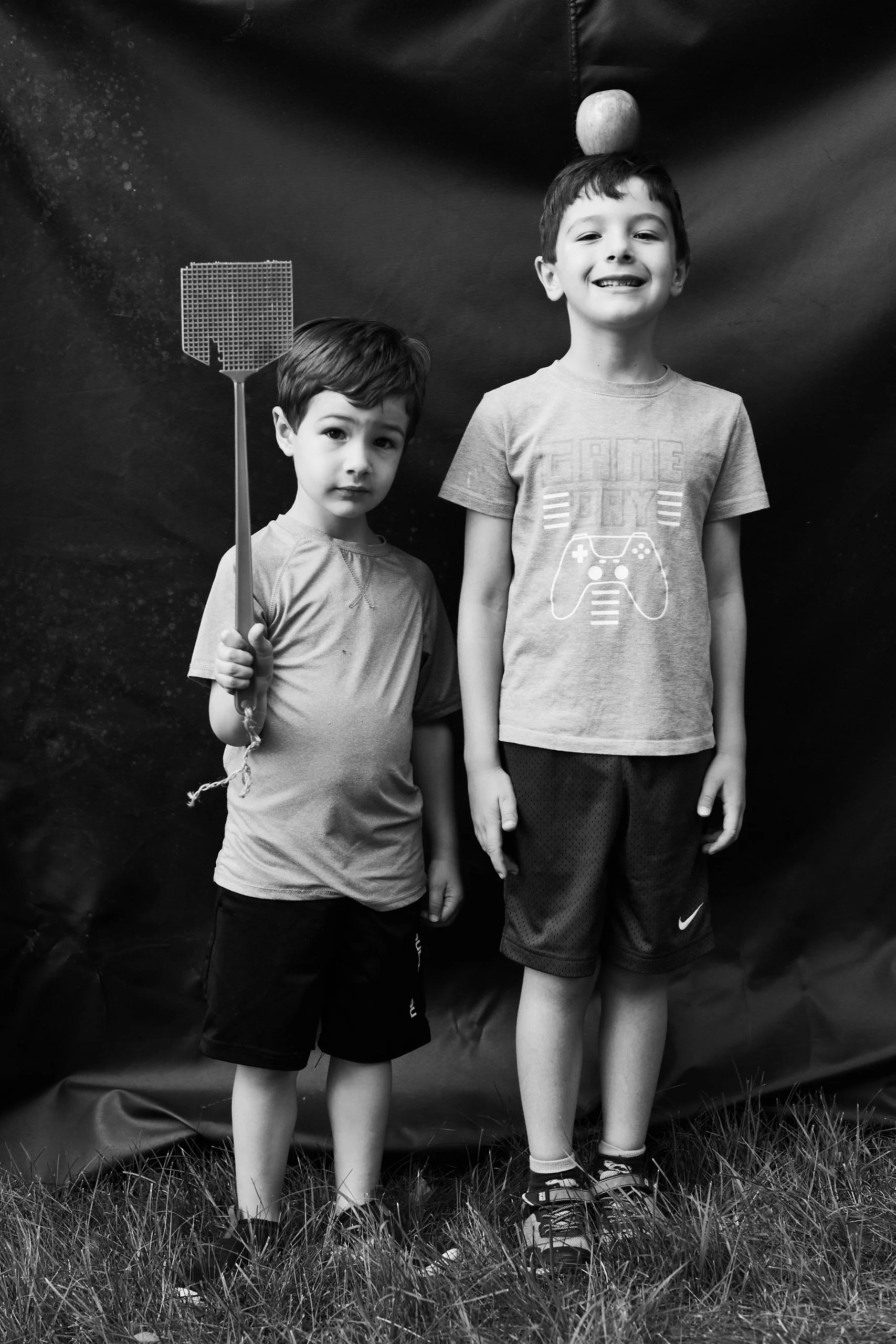 Two young boys standing outdoors on grass against a dark backdrop, one holding a fly swatter, the other balancing a ball on his head, both smiling.