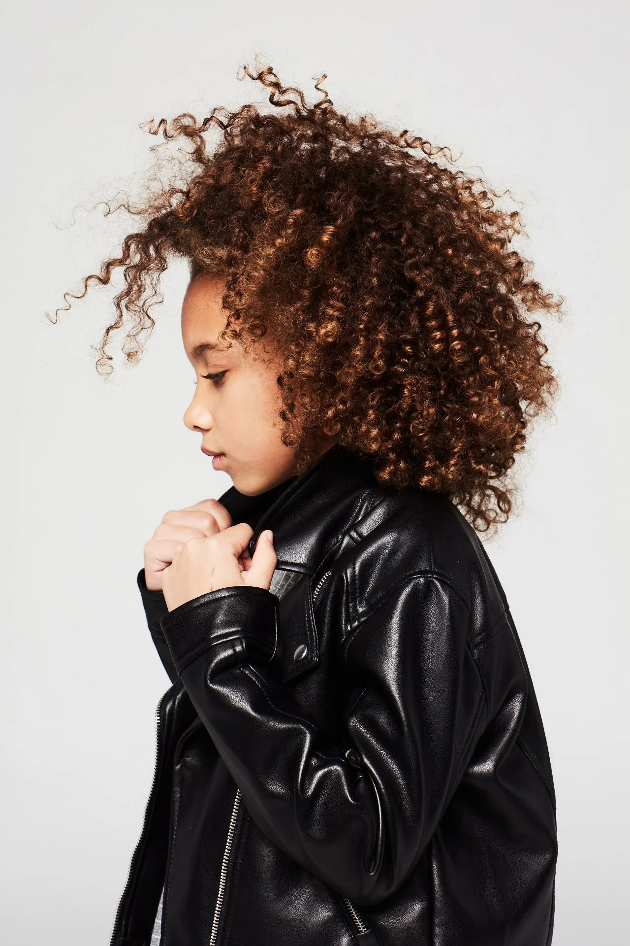 Side profile of a girl with curly brown hair wearing a black leather jacket against a plain grey background.