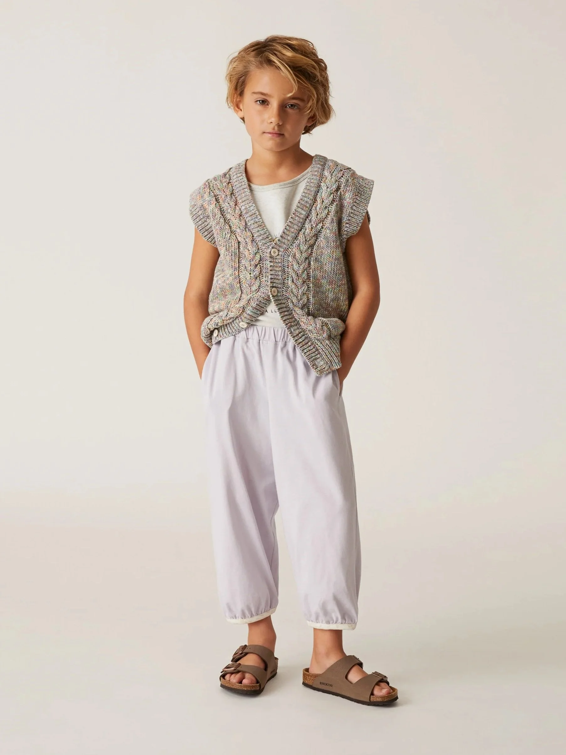 Young girl with blonde hair wearing a knit vest, white t-shirt, loose beige pants, and brown sandals.