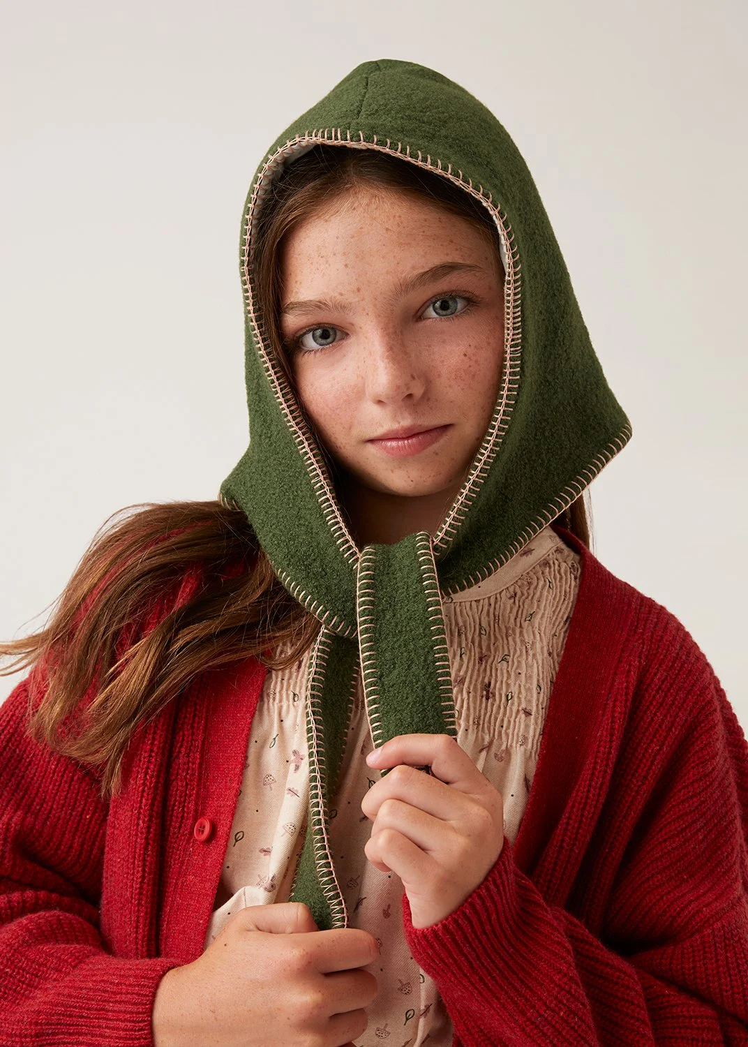 A young girl with light skin, freckles, and blue eyes, wearing a green hooded garment with pink stitching, a beige shirt with small patterns, and a red cardigan.