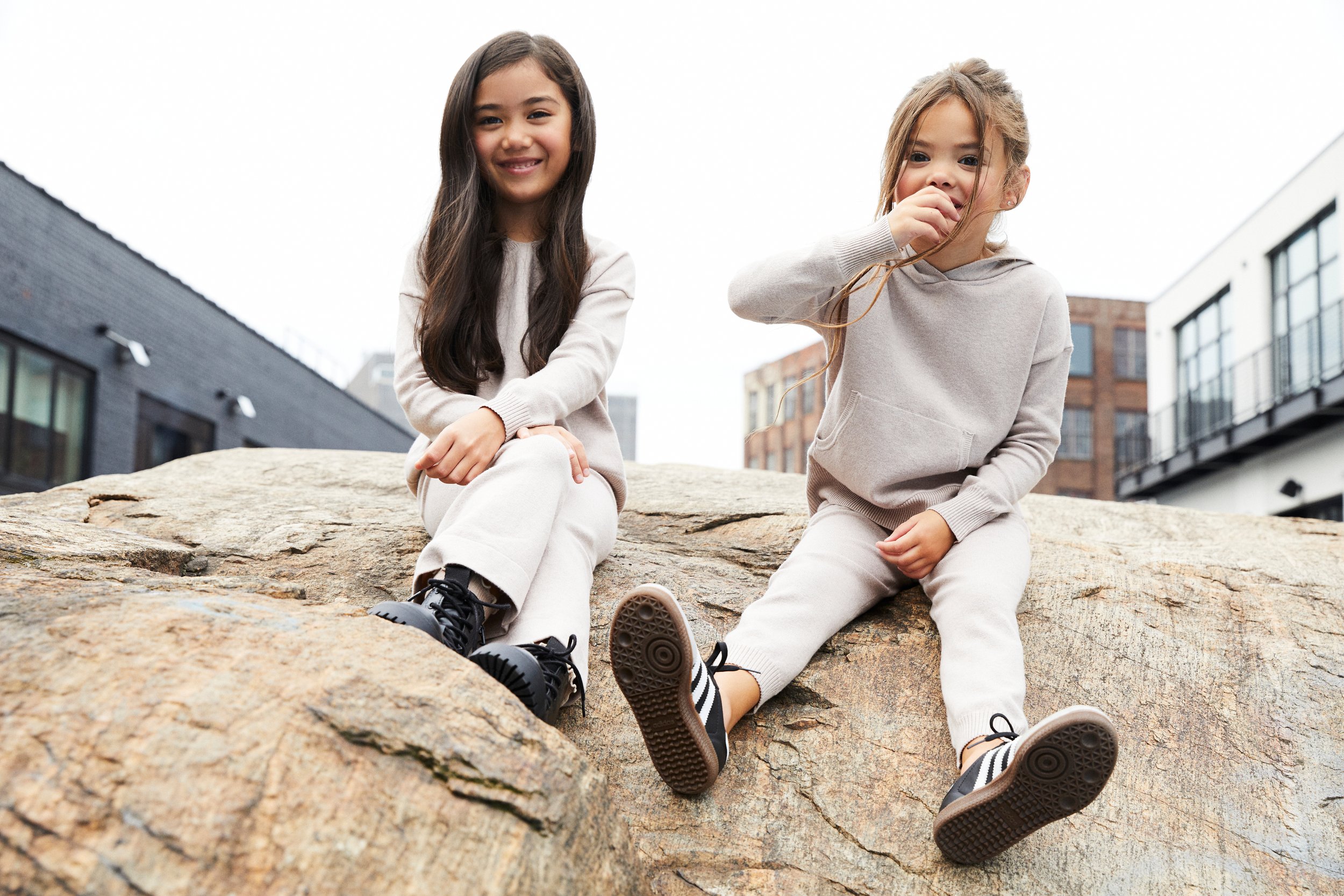 Two young girls sitting on a large rock outdoors, smiling and dressed in light-colored casual clothing.