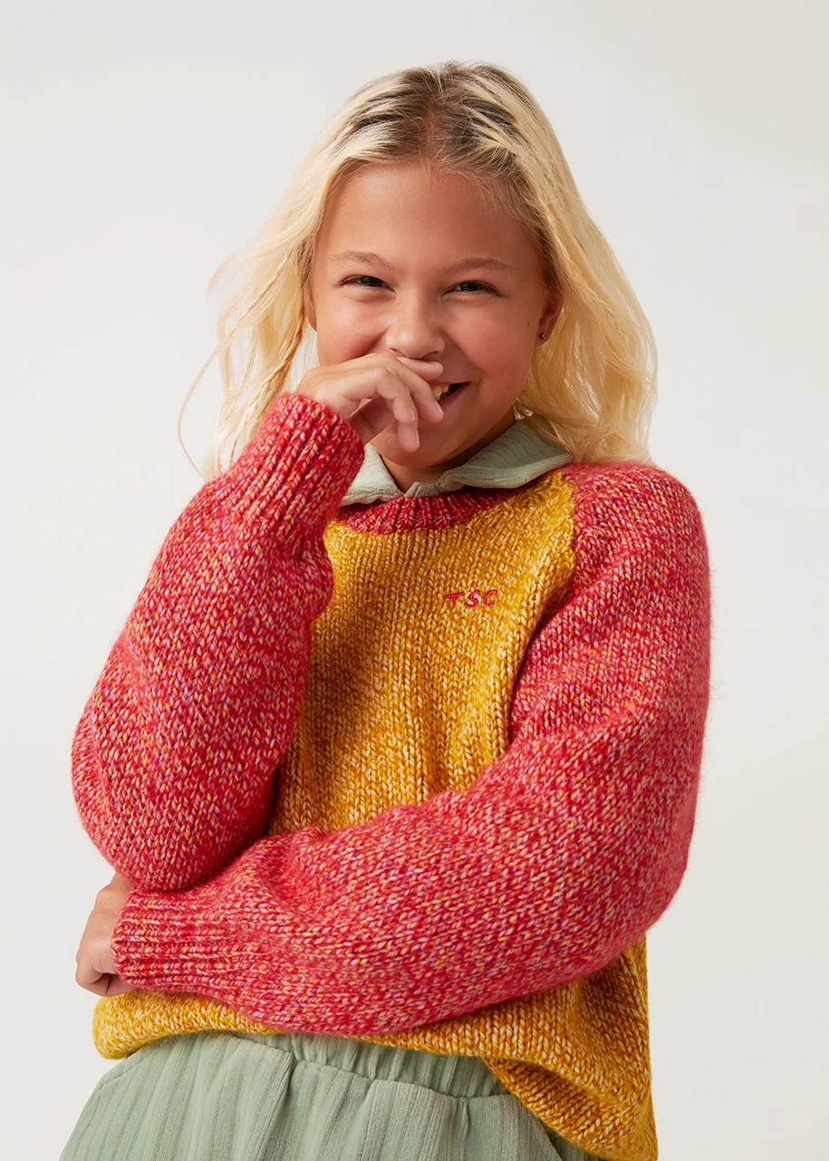A young woman with blonde hair, smiling and laughing while covering her mouth with her hand. She is wearing a red, yellow, and orange knit sweater over a light green shirt.