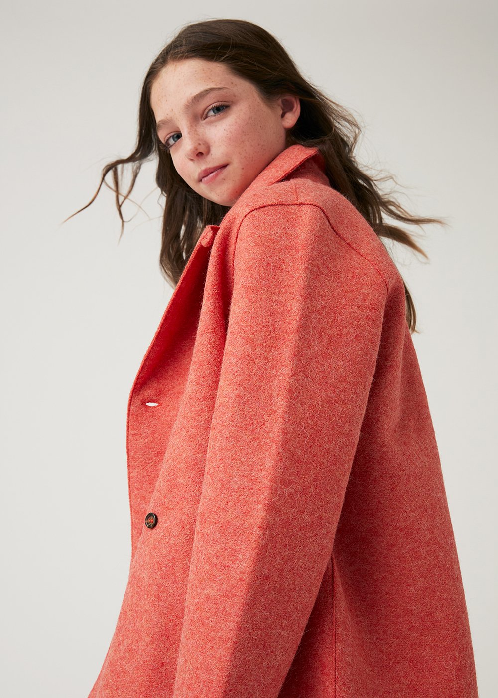 A young girl with long brown hair wearing a red coat, looking at the camera with a neutral expression.