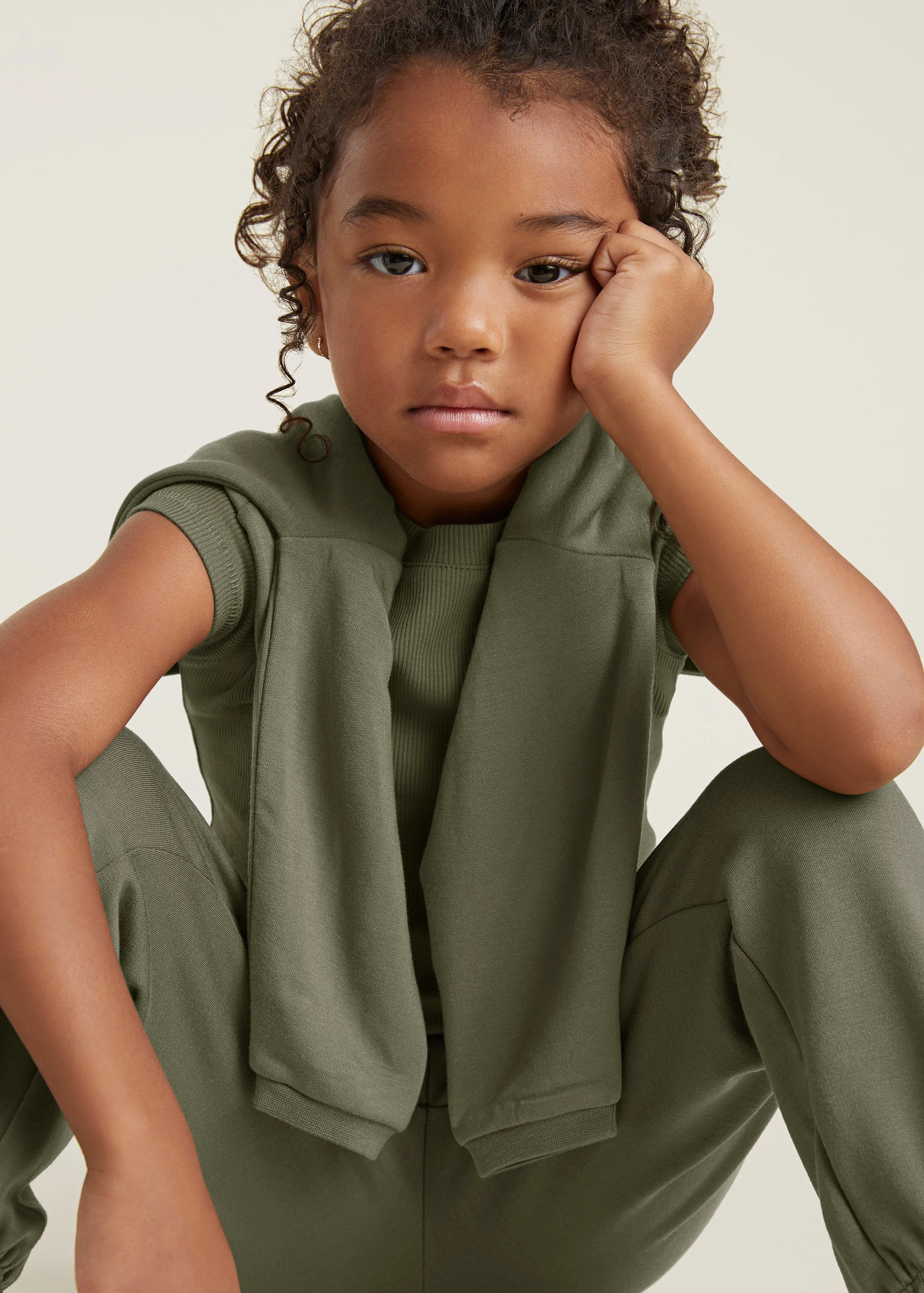 A young girl sitting with her knees up, resting her face on her hand, wearing an olive green t-shirt and matching pants, with a serious expression.