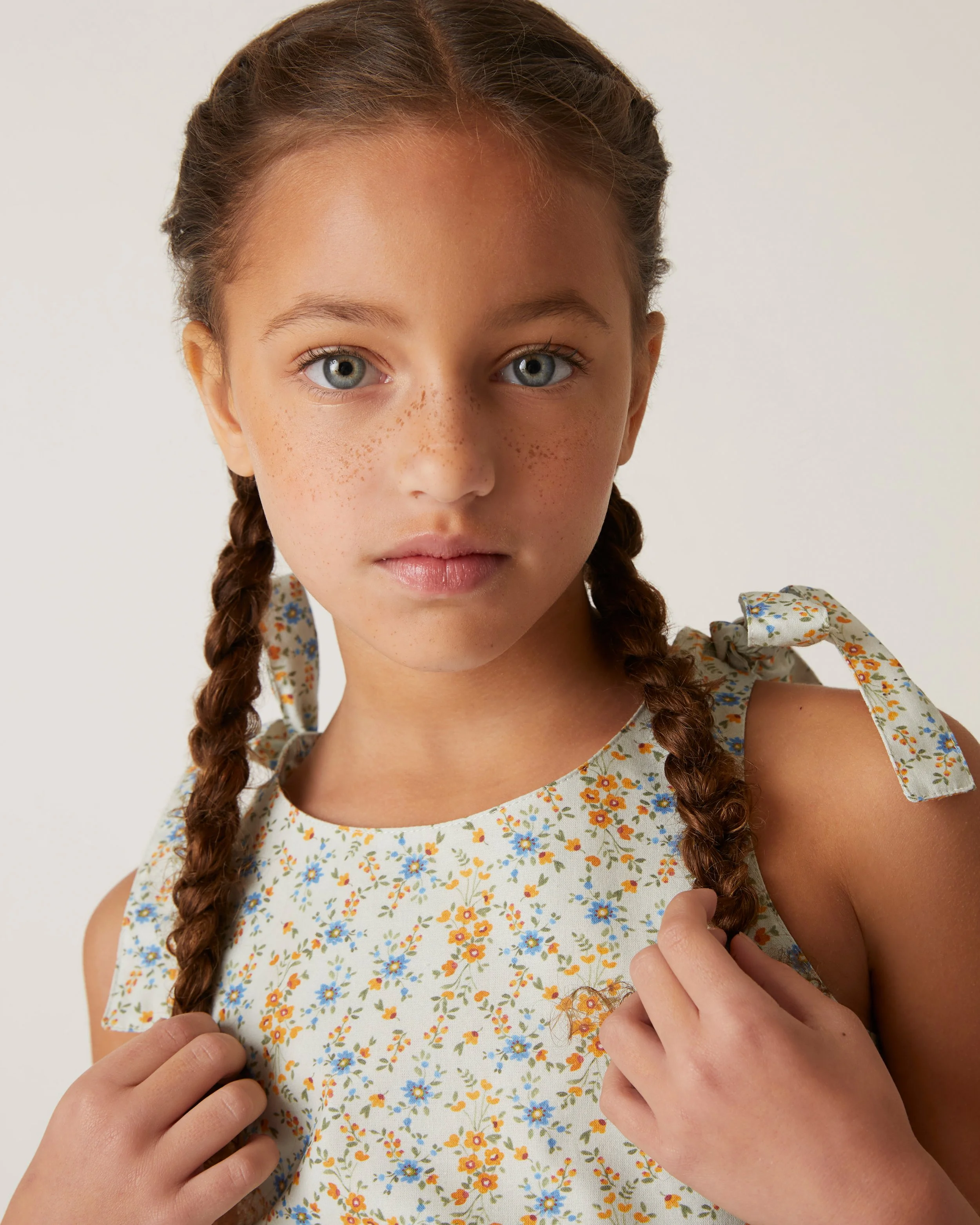 A young girl with blue eyes and freckles, wearing a cream-colored dress with a multicolored floral pattern and tied shoulder straps, holding her braided hair.