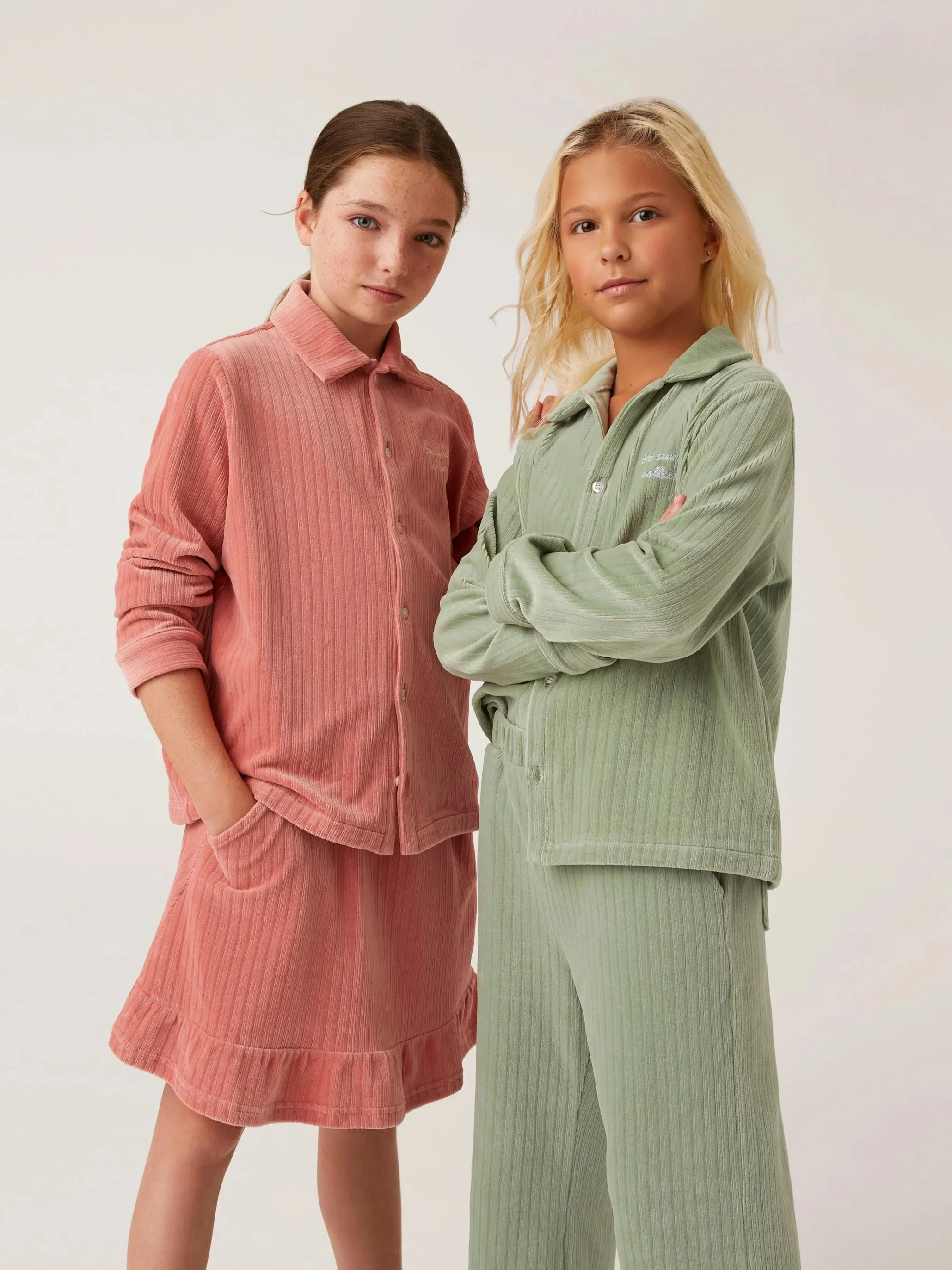 Two girls standing side by side, wearing pastel-colored pajamas, with one in pink and the other in light green, posing against a plain background.