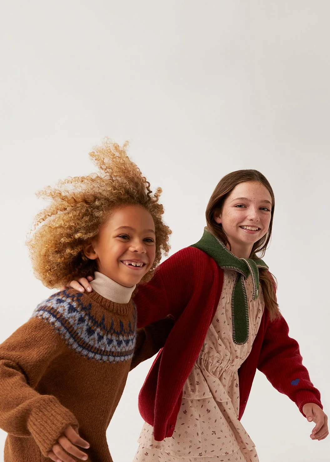 Two smiling girls, one with curly blonde hair wearing a brown patterned sweater, and the other with straight brown hair wearing a pink dress and red cardigan, are holding hands and posing together.