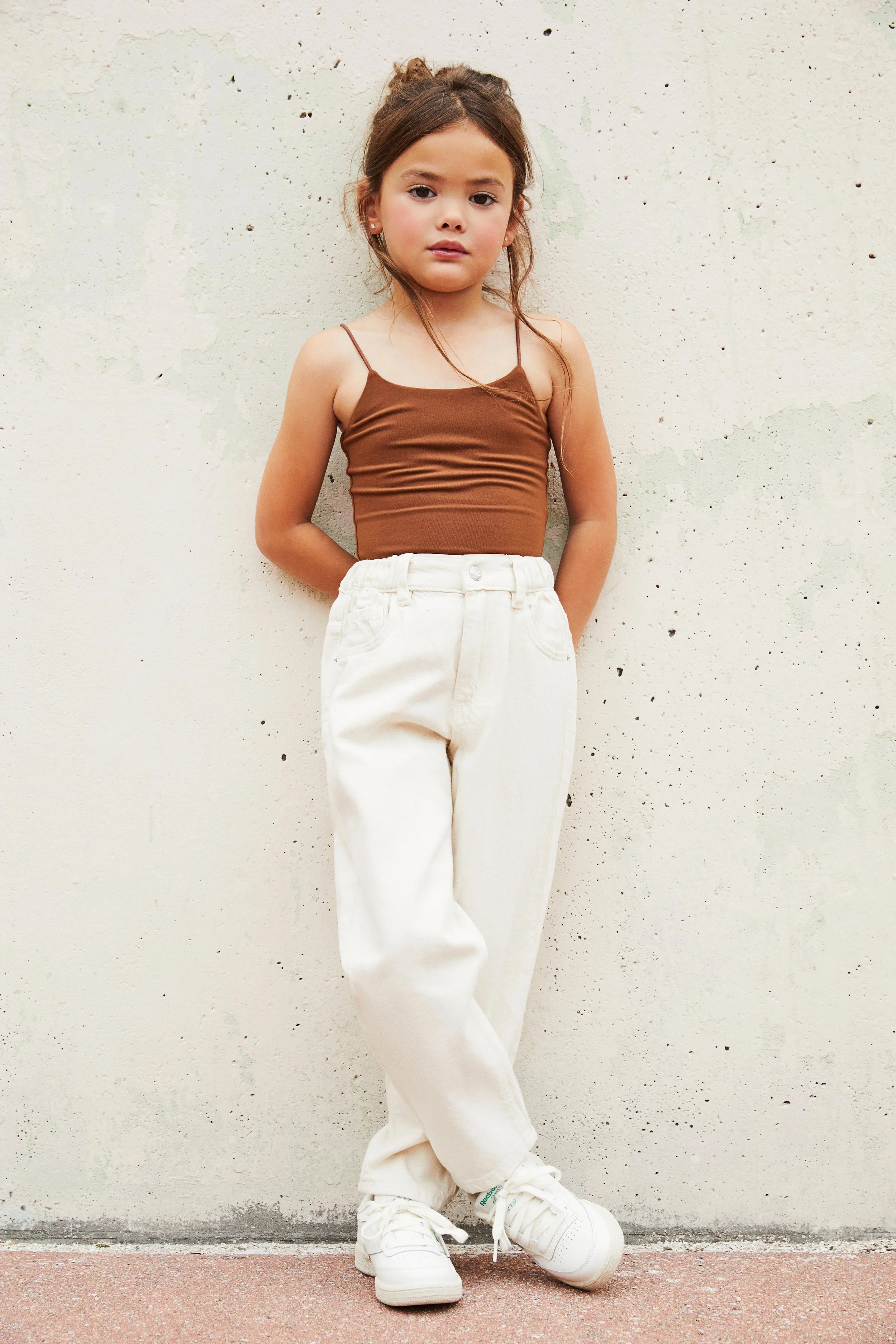 A young girl standing against a beige wall, wearing a brown spaghetti strap top, cream-colored pants, and white sneakers, with her hands behind her back.