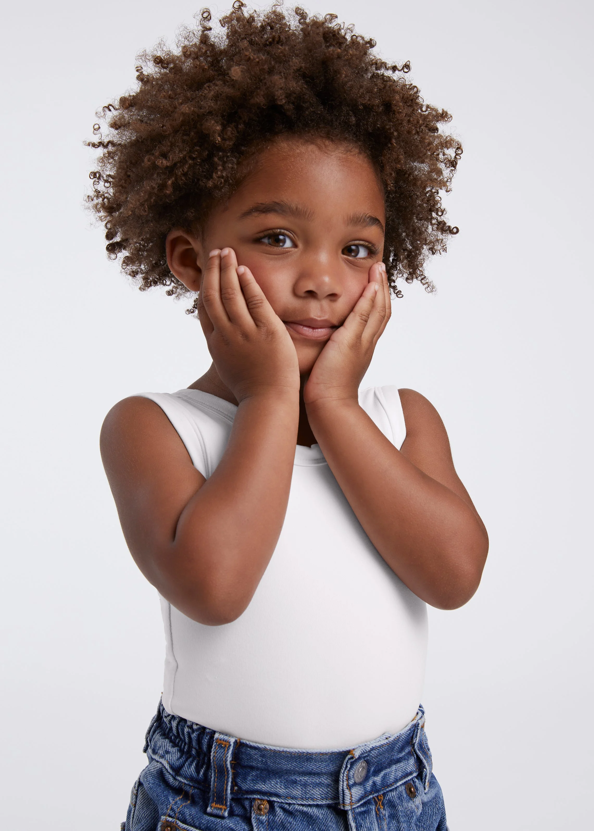 A young girl with curly hair, wearing a white sleeveless top and blue denim shorts, holding her face with both hands and looking at the camera with a gentle smile.