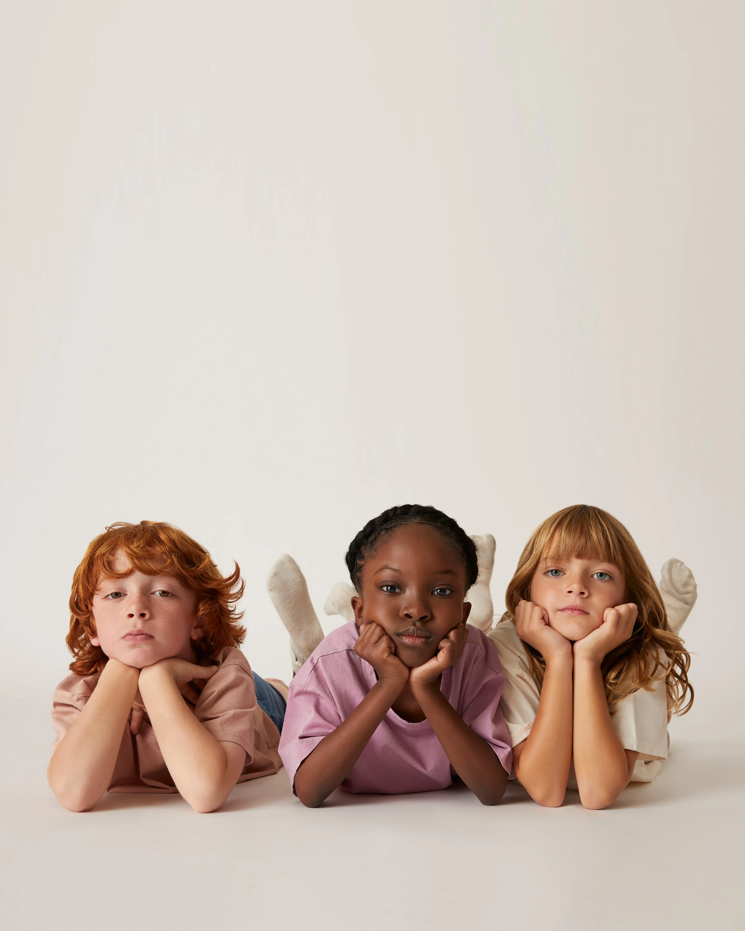 Three children lying on the floor with chins resting on their hands, looking at the camera with neutral expressions, against a plain white background.
