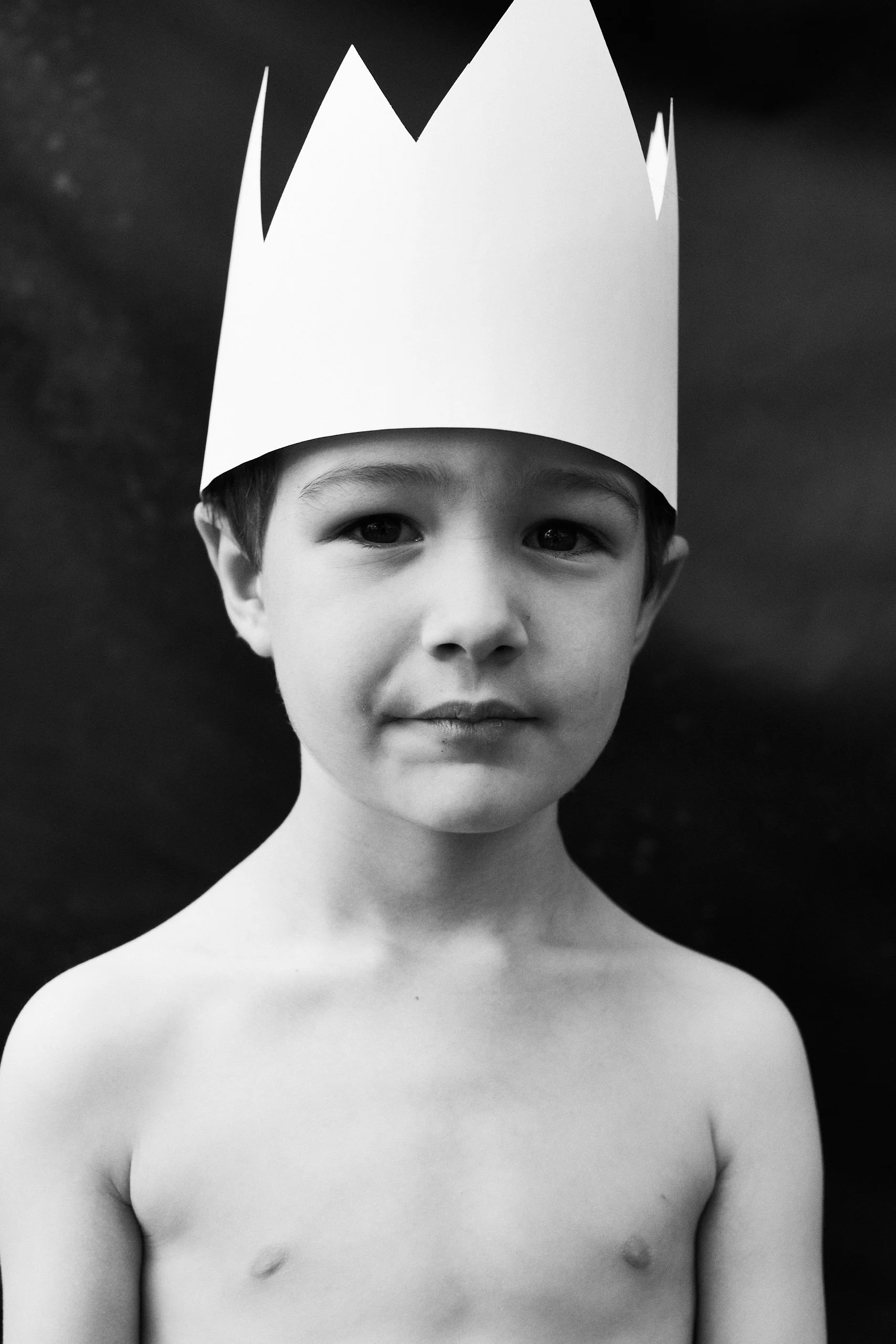 A young shirtless boy wearing a paper crown against a dark background.