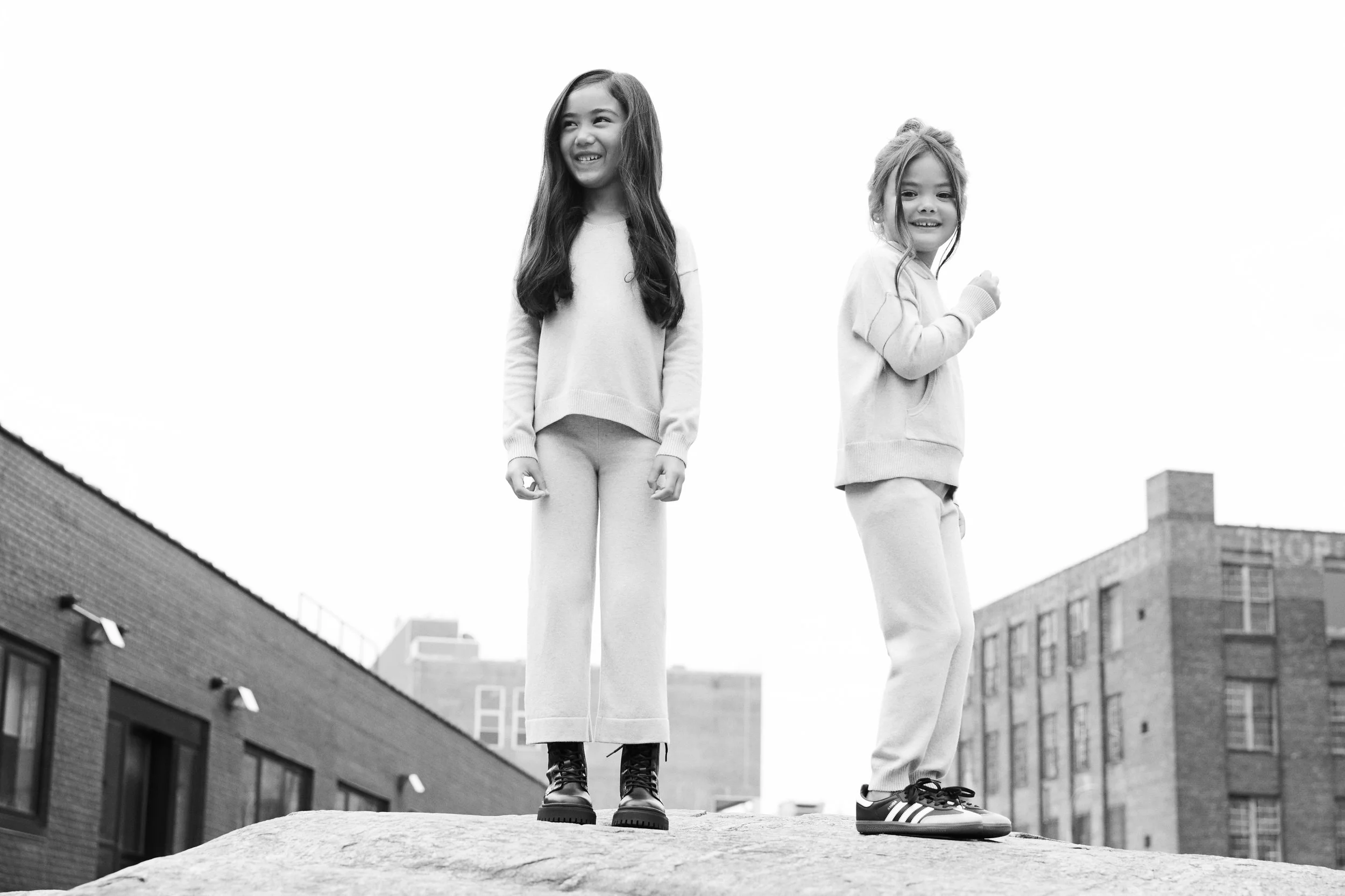 Two young girls standing on a ledge outdoors, smiling and looking off into the distance, with buildings in the background.