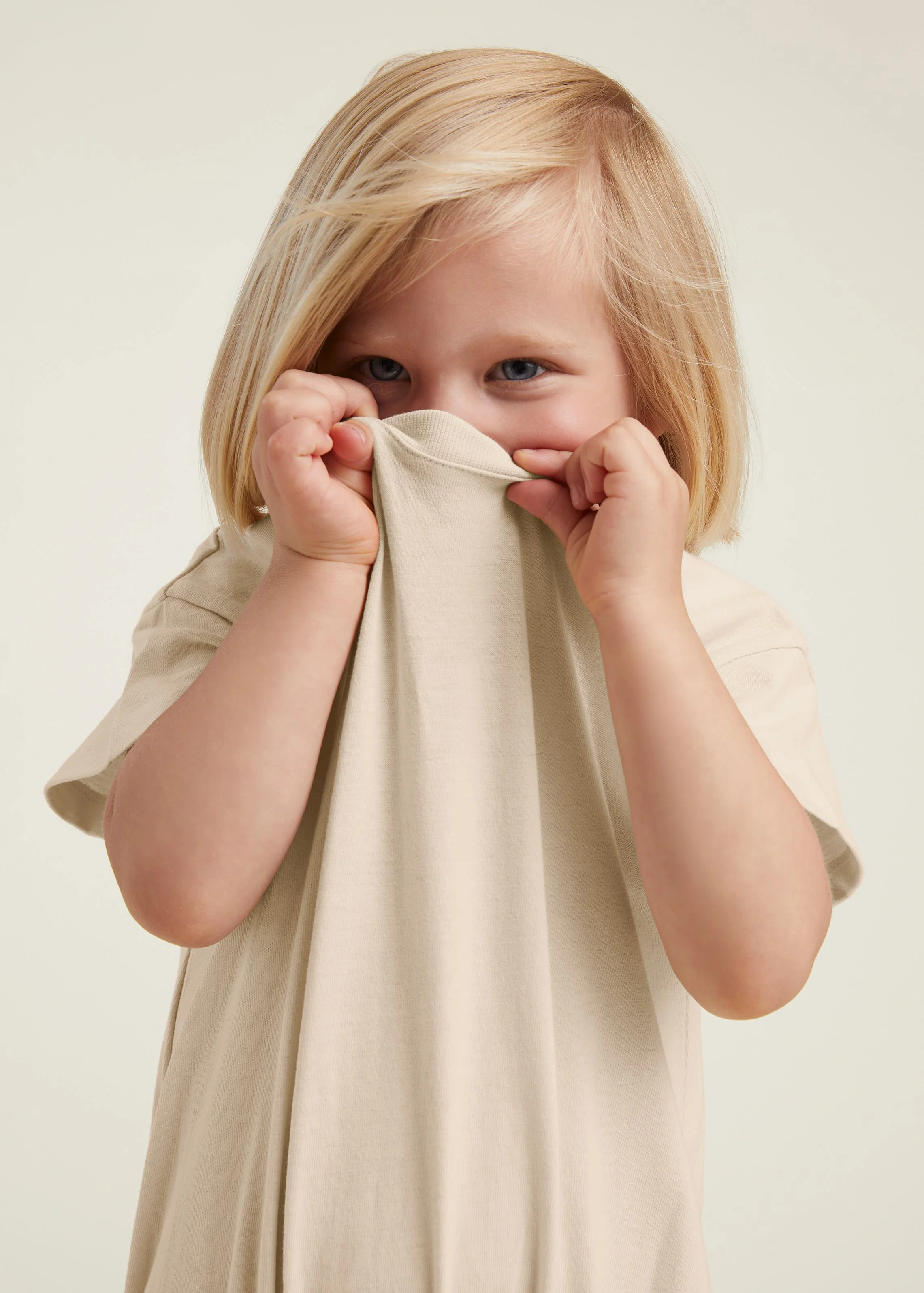 A young girl with blonde hair wearing a beige shirt, holding it up to her nose and smiling.