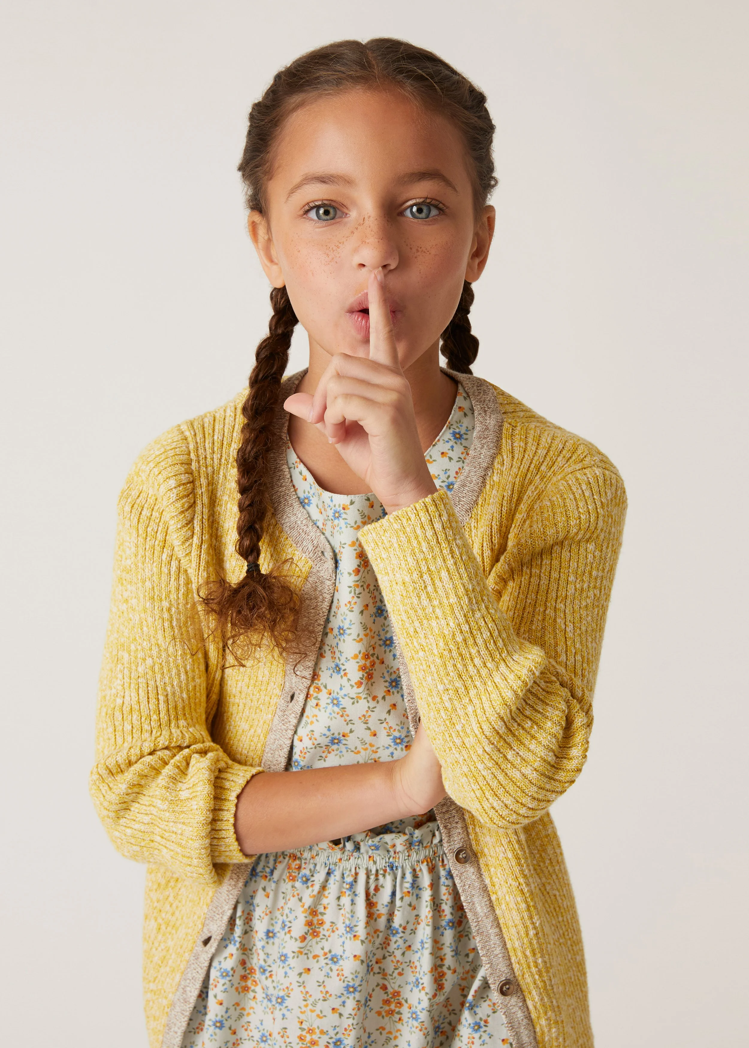 A young girl with braided hair, blue eyes, and freckles holding her finger to her lips in a 'shh' gesture, wearing a yellow cardigan over a floral dress.