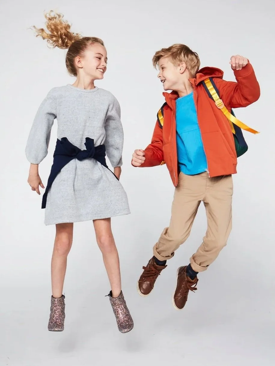 A girl and boy are smiling and jumping in the air, facing each other. The girl has curly blonde hair and is wearing a gray dress with a dark bow around her waist, and sparkly boots. The boy has blonde hair, and is wearing a blue shirt, an orange jacket, khaki pants, and brown shoes, with a backpack on his back.