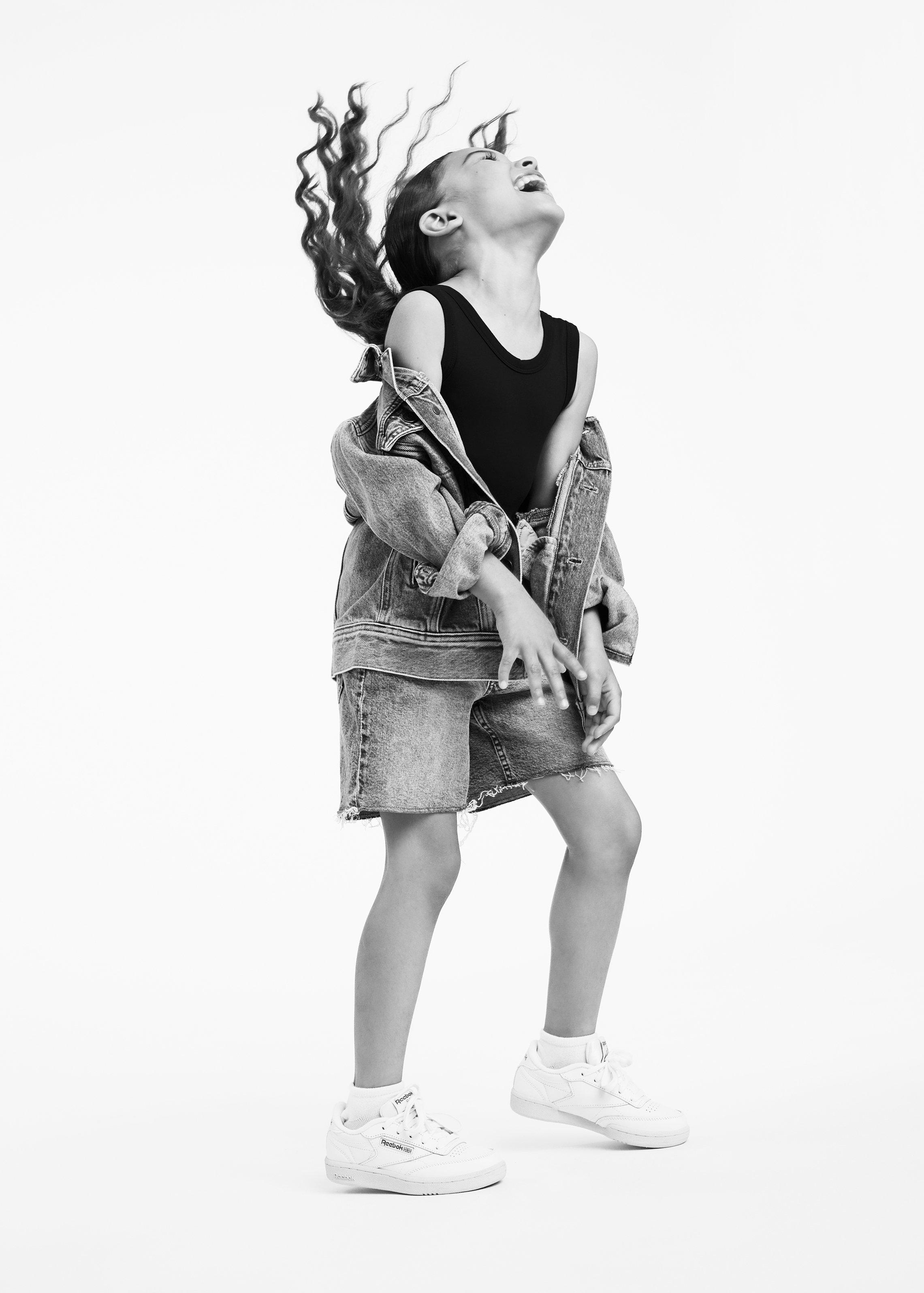 A person with long hair in motion, wearing a black sleeveless top, denim jacket and skirt, and white sneakers, standing against a plain background.