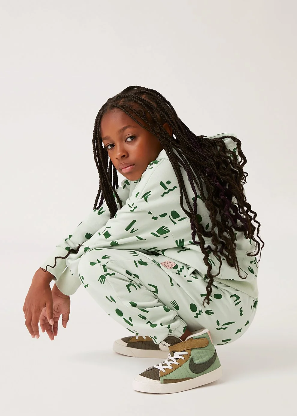 A young girl squatting in a white studio background, wearing a green and white patterned tracksuit and sneakers, with long braided hair.