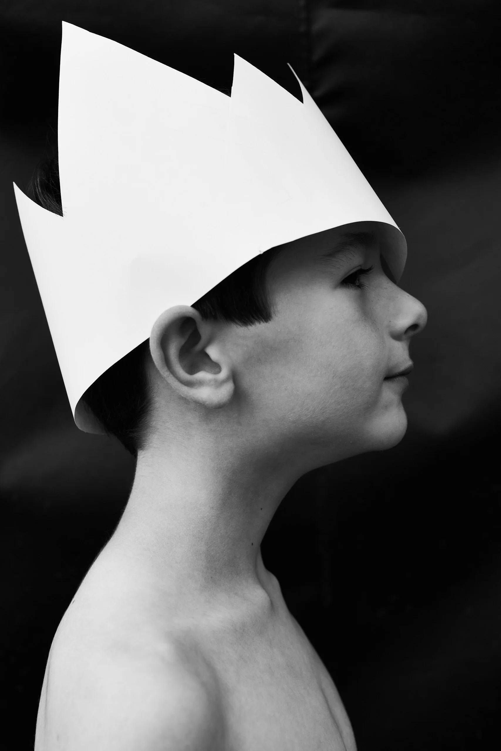 A young boy in profile, wearing a paper crown, black and white photograph.