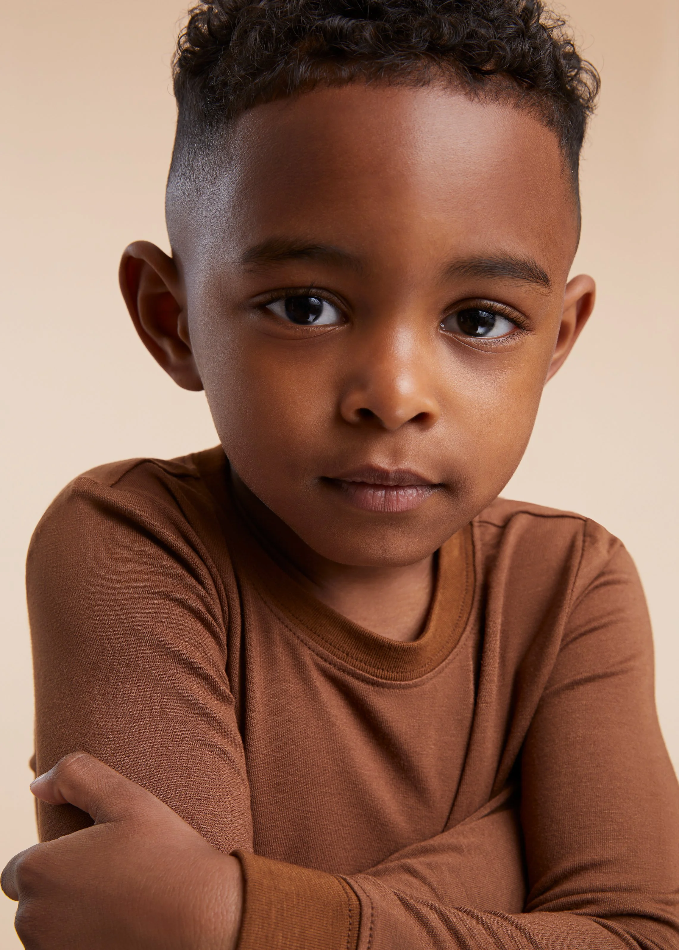 Close-up portrait of a young boy with dark skin, short curly hair, wearing a brown long-sleeve shirt, looking directly at the camera with a calm expression.